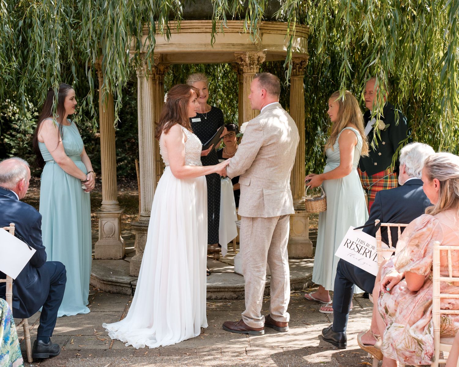 Outdoor wedding ceremony under willow tree with bride in white dress and groom in tan suit exchanging vows.