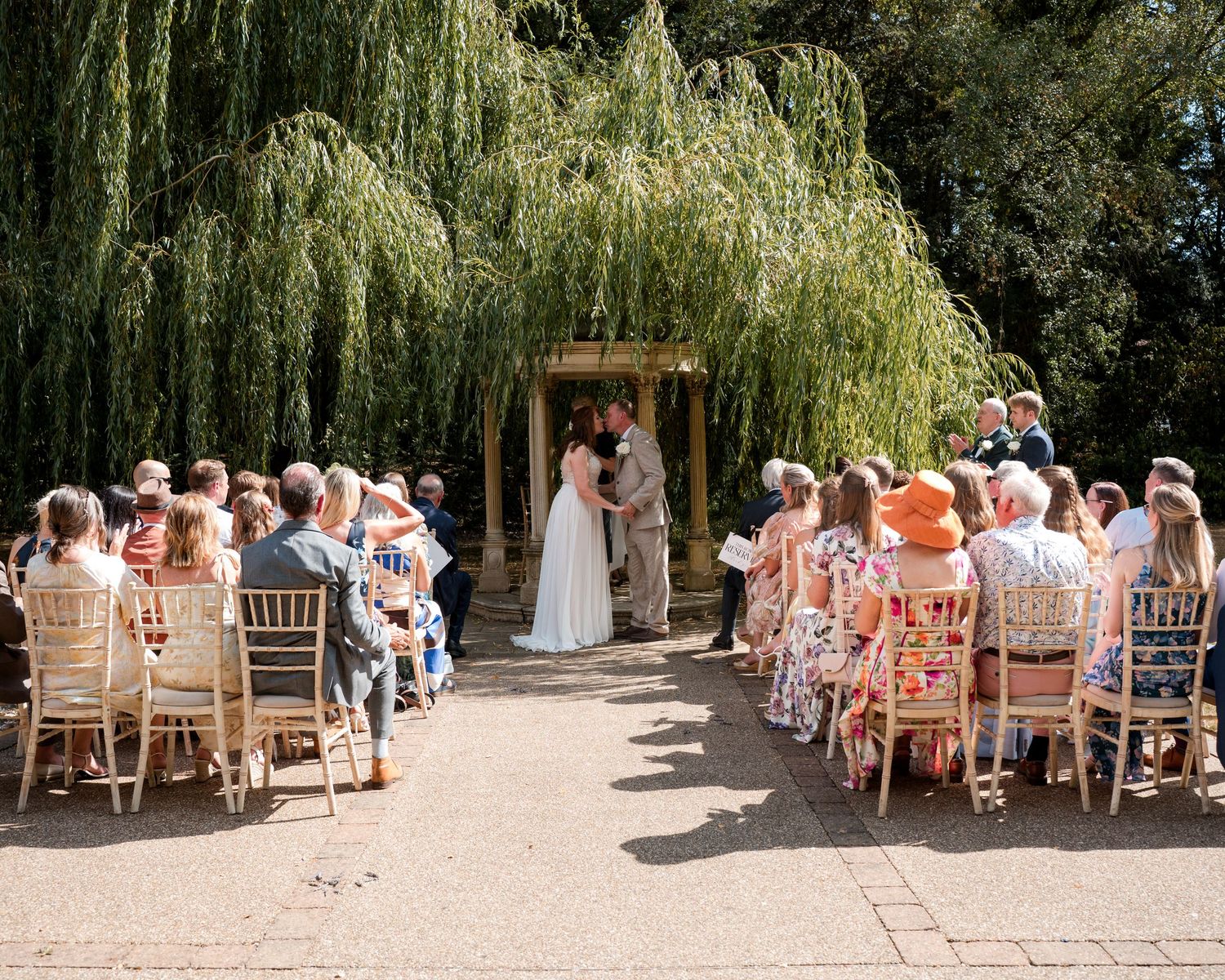 Outdoor wedding ceremony under a weeping willow tree with guests seated in wooden chairs on a sunny day.