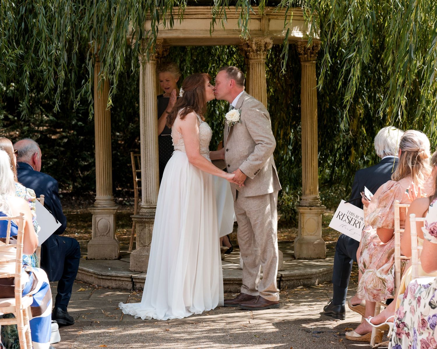Bride and groom share first kiss at outdoor wedding ceremony under willow tree with stone gazebo.