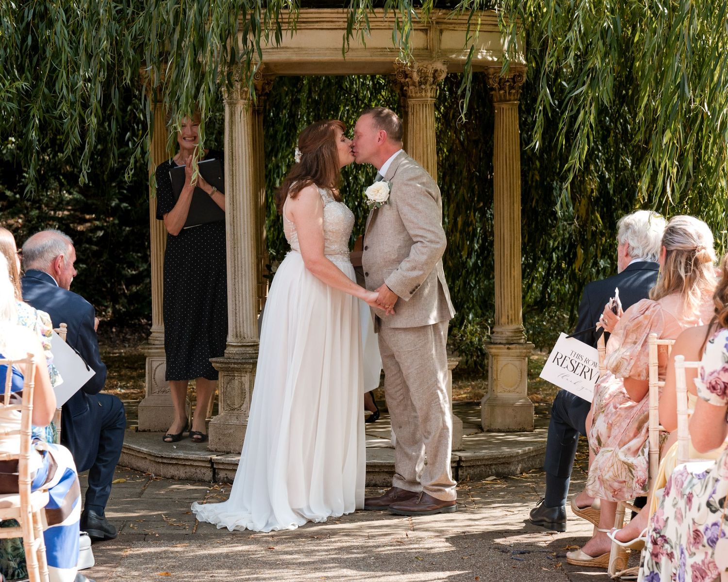 Bride and groom share first kiss at outdoor wedding ceremony under willow tree with guests seated alongside.