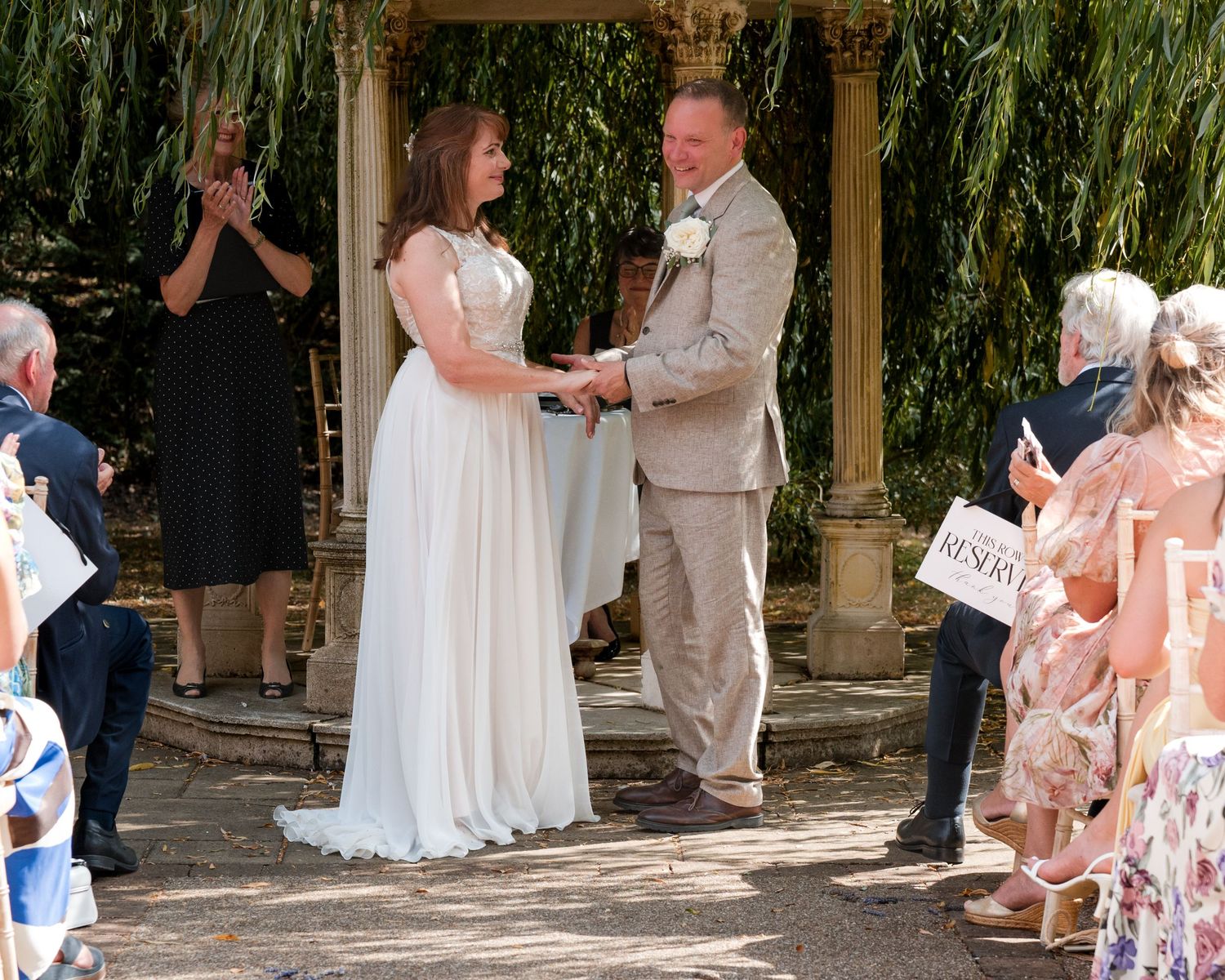 Bride and groom exchange vows at outdoor wedding ceremony under willow tree with guests watching.