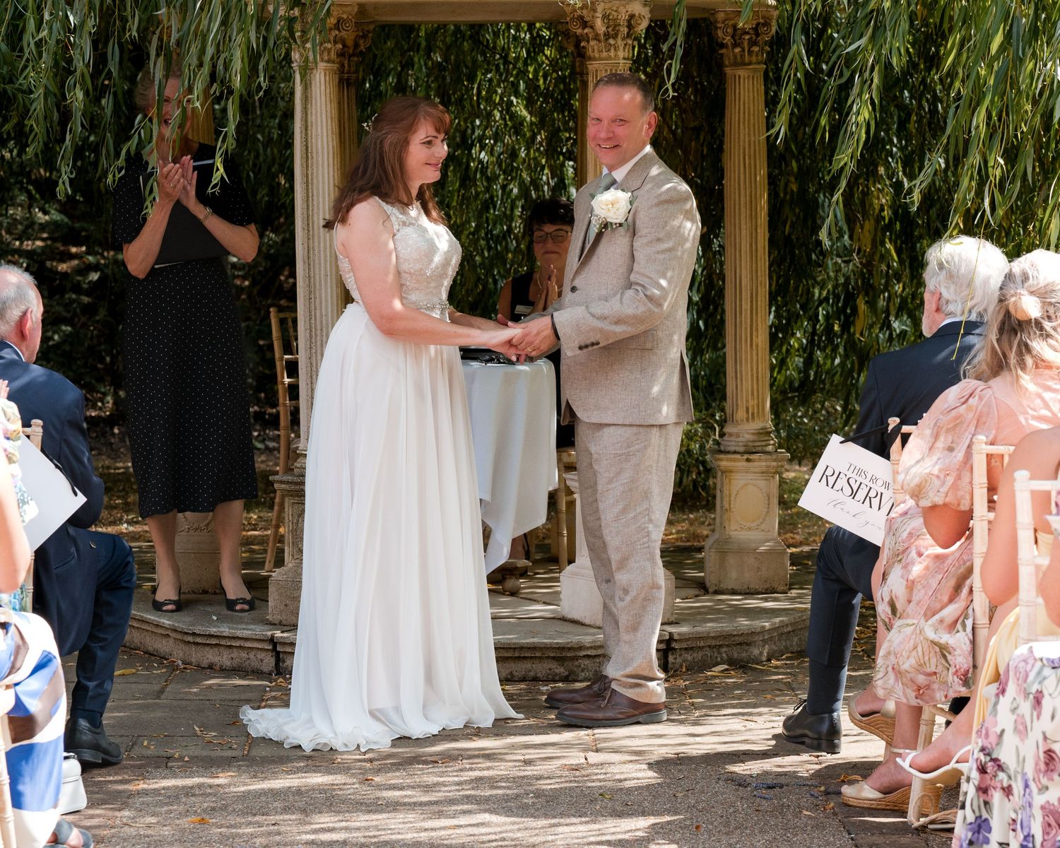 Bride and groom holding hands during outdoor wedding ceremony under willow tree with guests applauding.