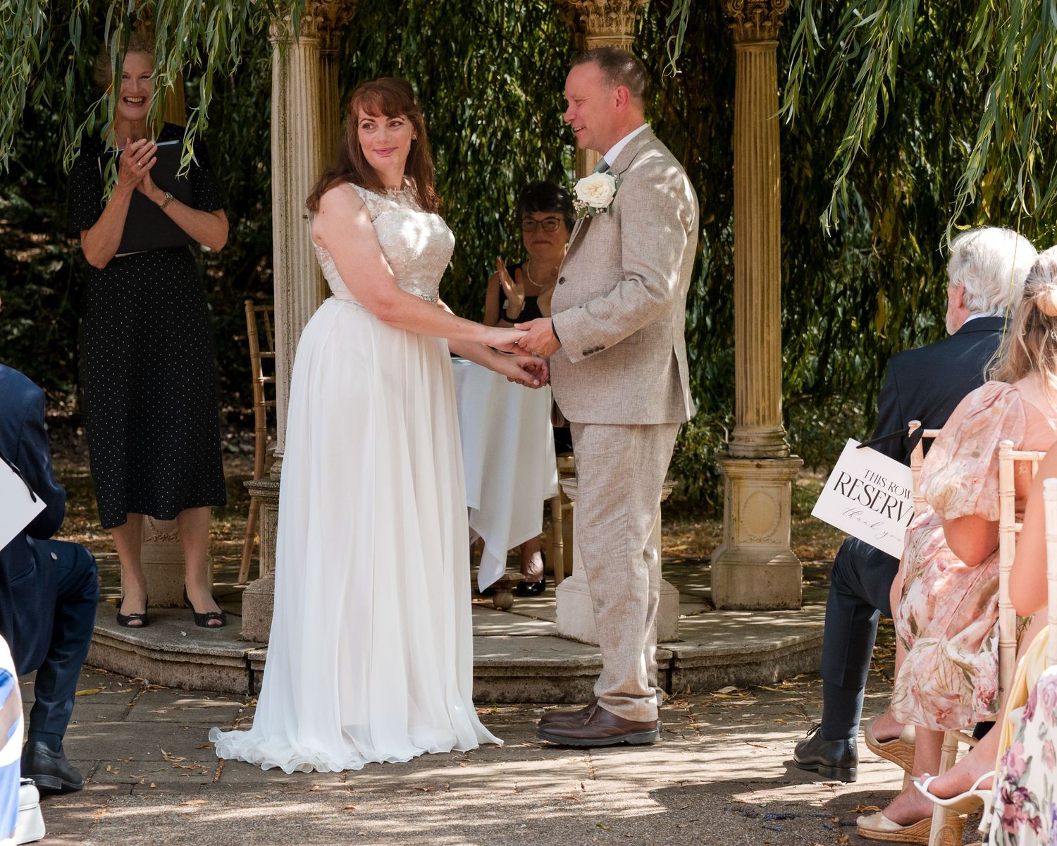 Bride and groom exchange vows outdoors under a willow tree, surrounded by guests at a sunny wedding ceremony.