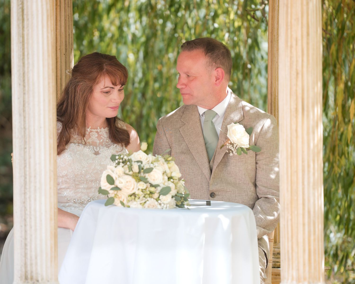 Bride and groom share an intimate moment at outdoor wedding ceremony, bride holding white bouquet near elegant pillars.