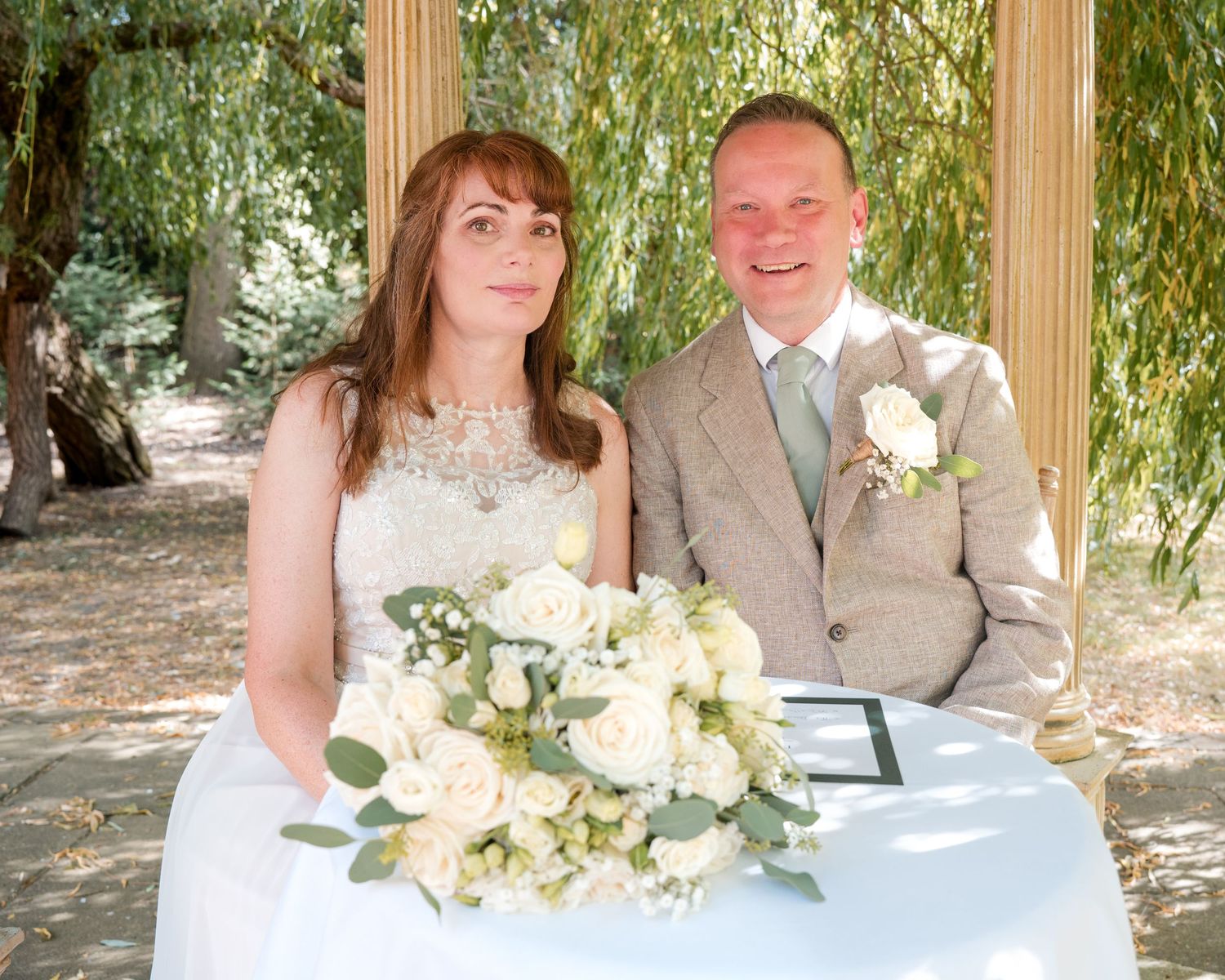 Smiling bride and groom sitting together at outdoor wedding ceremony table with white rose bouquet.