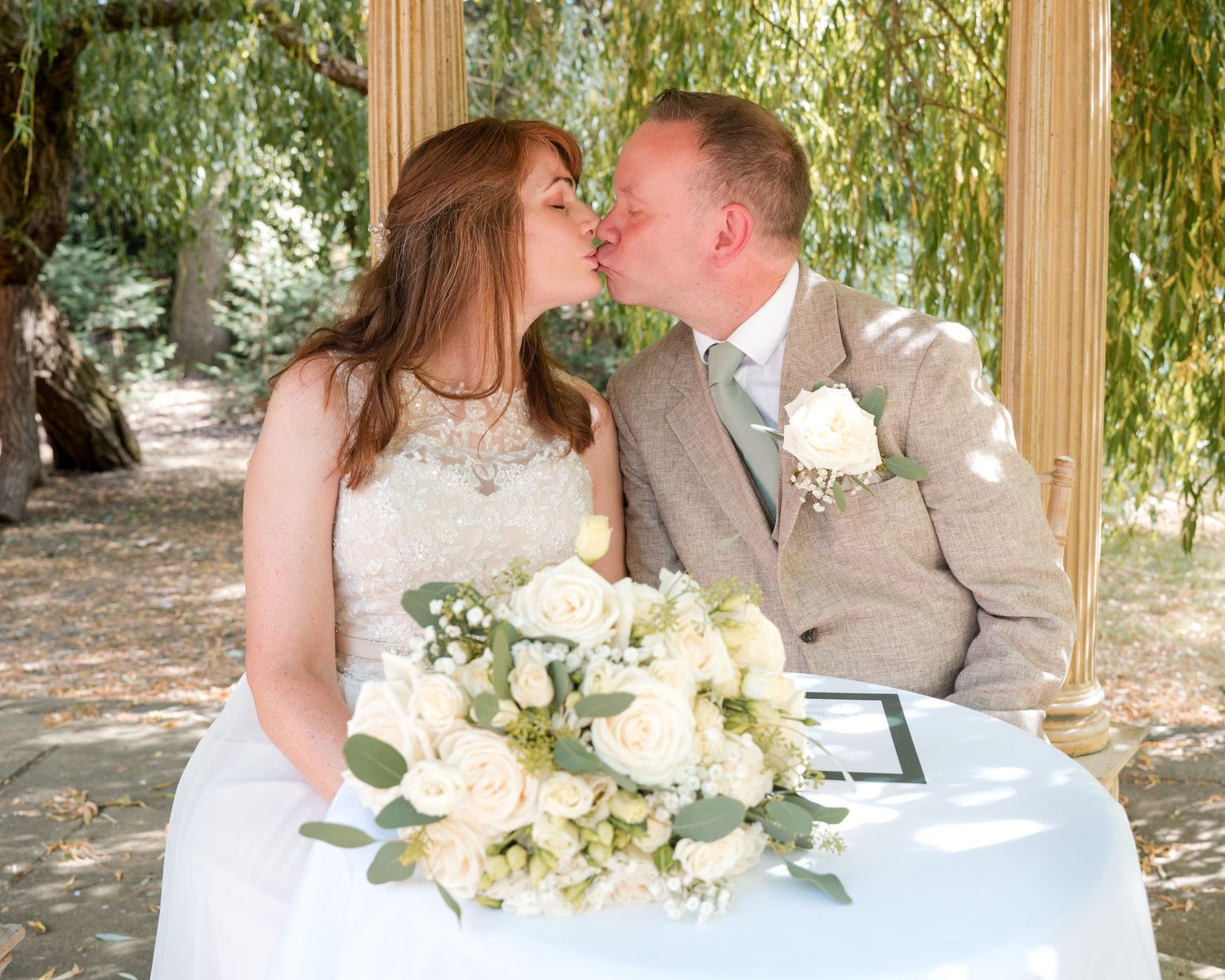 Bride and groom share a kiss at outdoor wedding ceremony, white rose bouquet on table, beneath weeping willow tree.