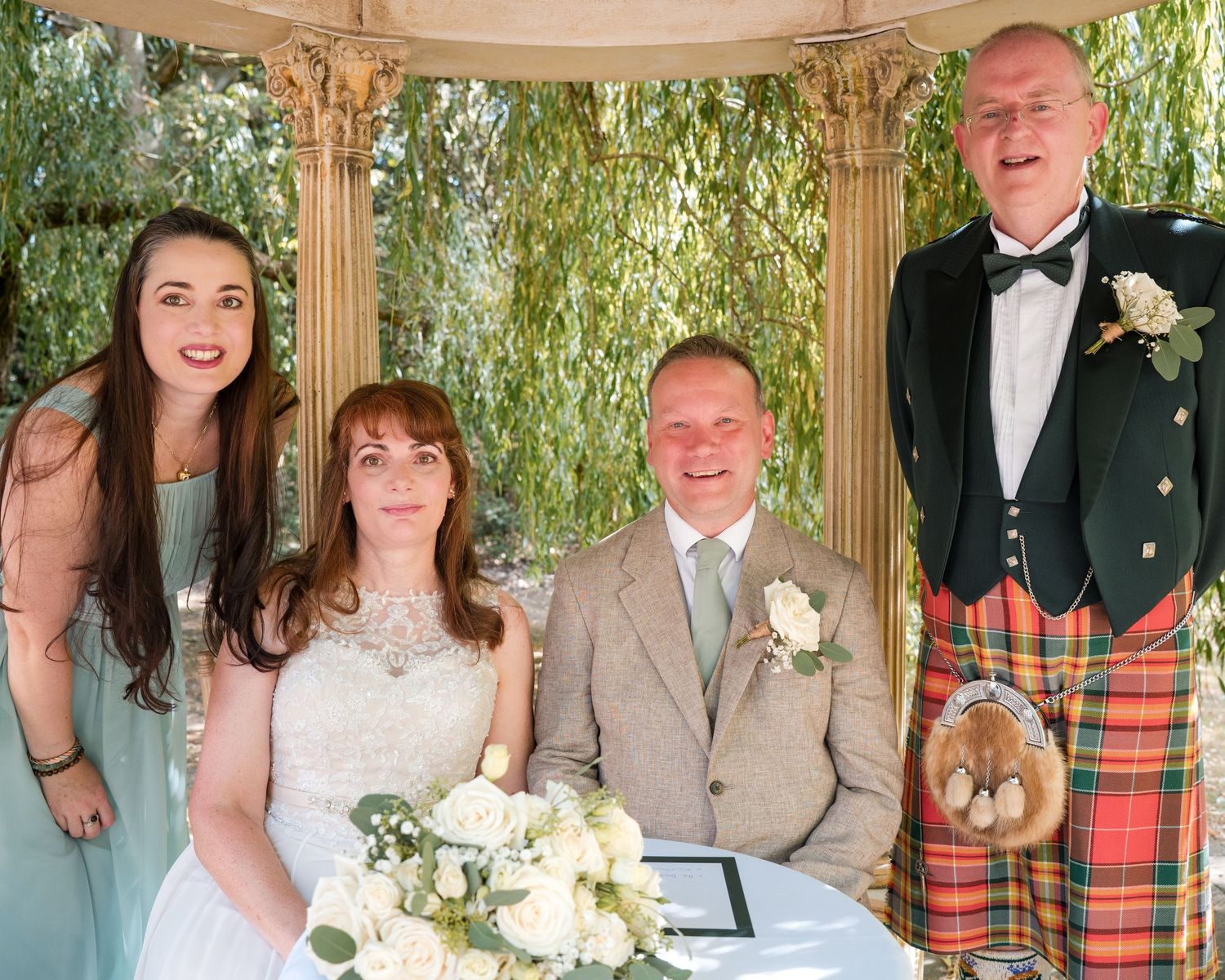Wedding party posing under floral gazebo, bride in white gown, groom in kilt, bridesmaid in mint dress.