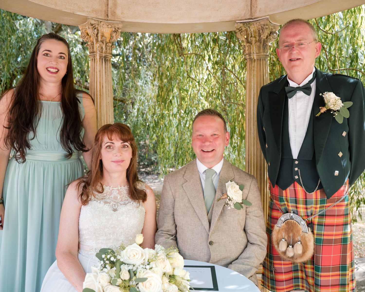 Wedding couple signs register seated under gazebo, flanked by bridesmaid in mint dress and man in Scottish tartan kilt.