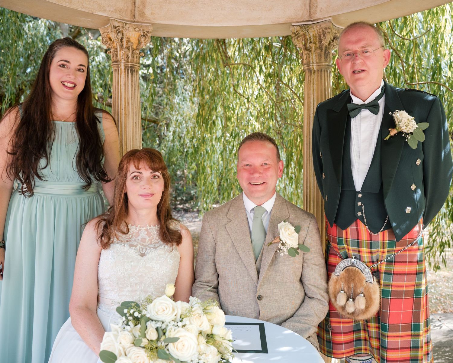 Wedding couple seated with bridesmaid and kilted groomsman under floral gazebo outdoors.