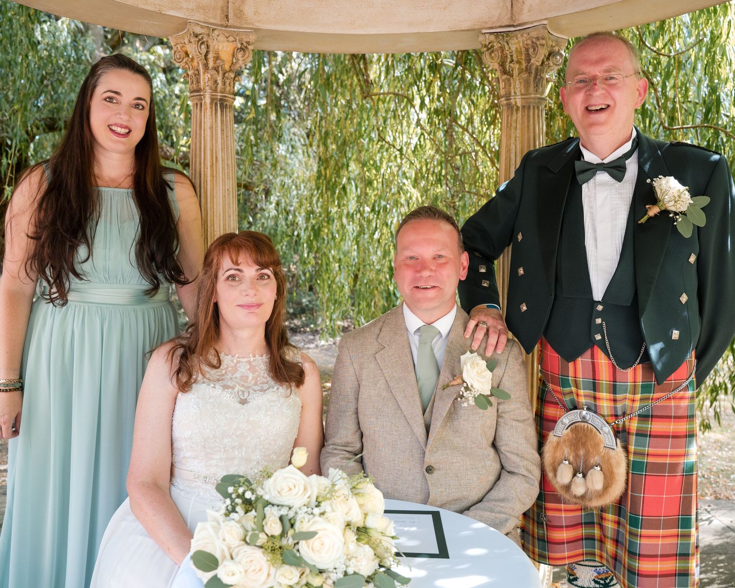 Wedding party posing under gazebo, bride in white gown, groom in kilt, bridesmaid in mint dress, outdoor ceremony.