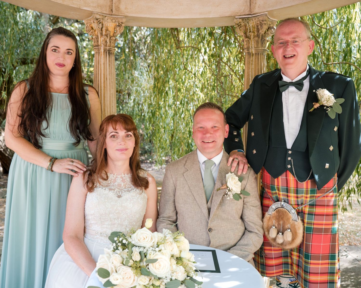 Wedding party poses under gazebo, bride and groom seated, bridesmaid and kilted man standing behind them.