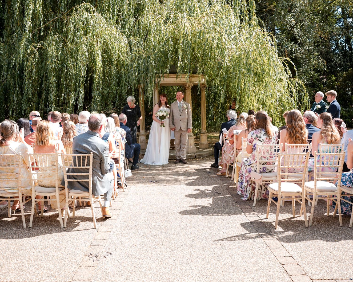 Outdoor wedding ceremony under a willow tree with guests seated in wooden chairs along an aisle leading to the couple.