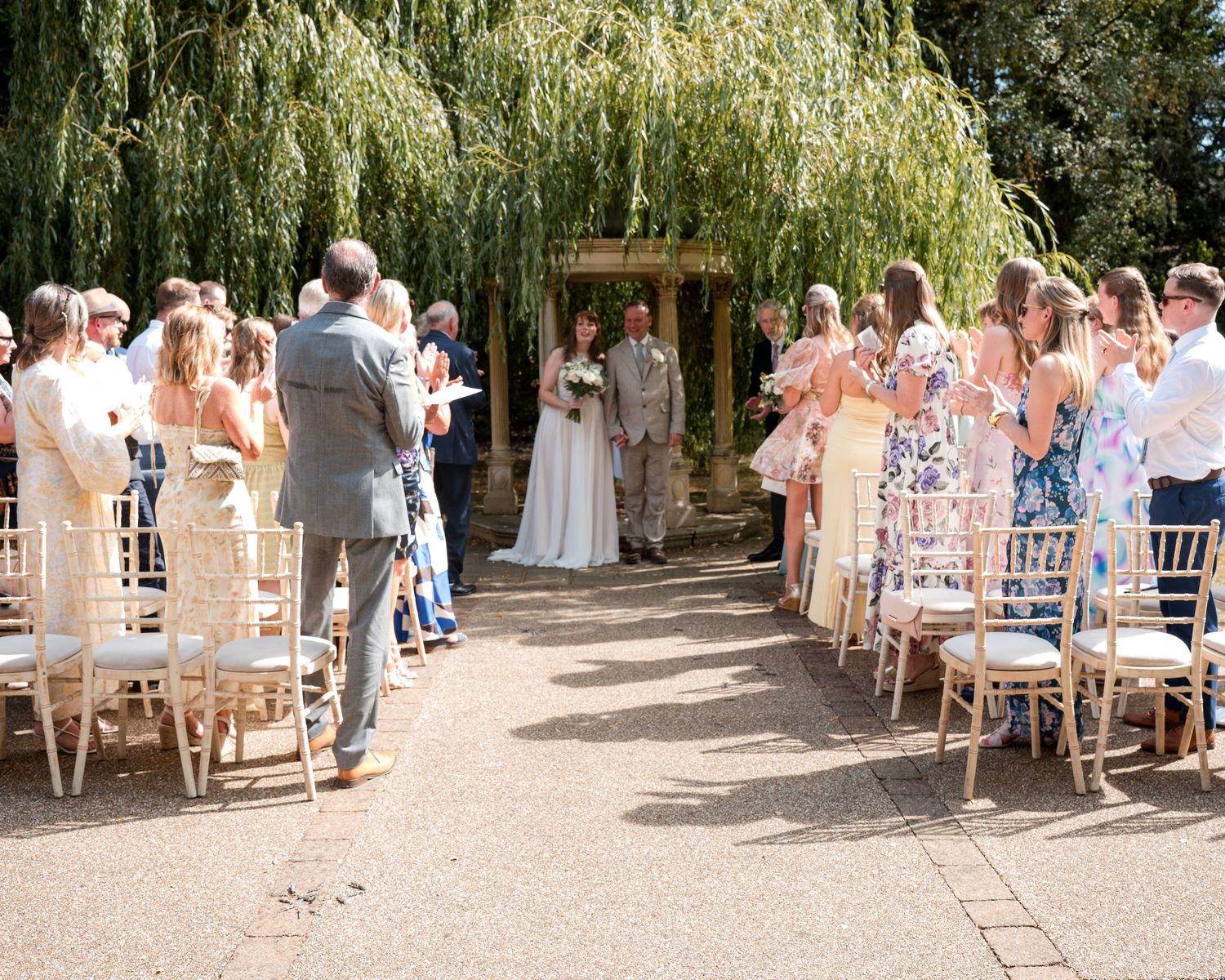 Outdoor wedding ceremony with guests seated in gold chairs along an aisle leading to a couple under a willow tree.
