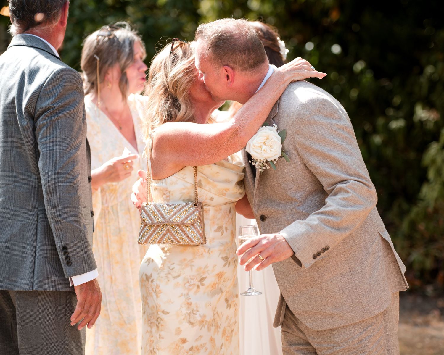 Bride and groom share a kiss at outdoor wedding ceremony, bride in floral dress, groom in tan suit with white boutonniere.
