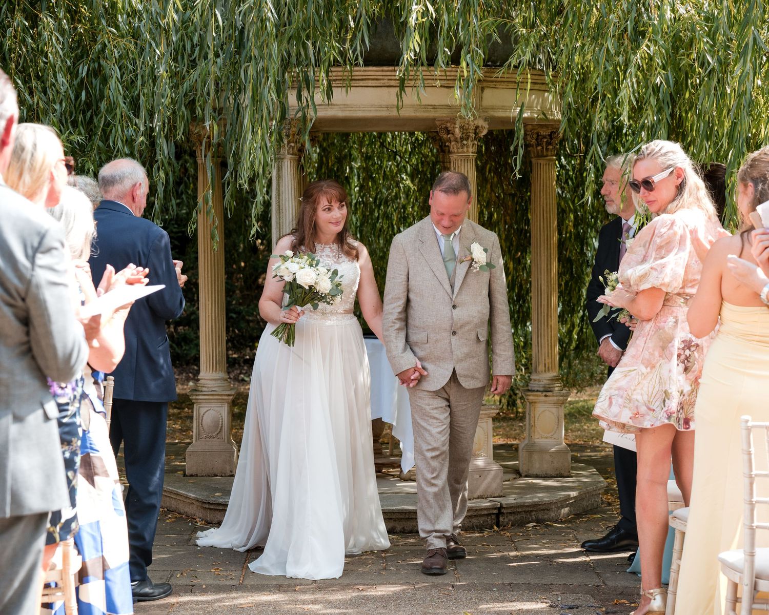 Bride and groom walk down the aisle outdoors under weeping willow trees at a sunny garden wedding ceremony.