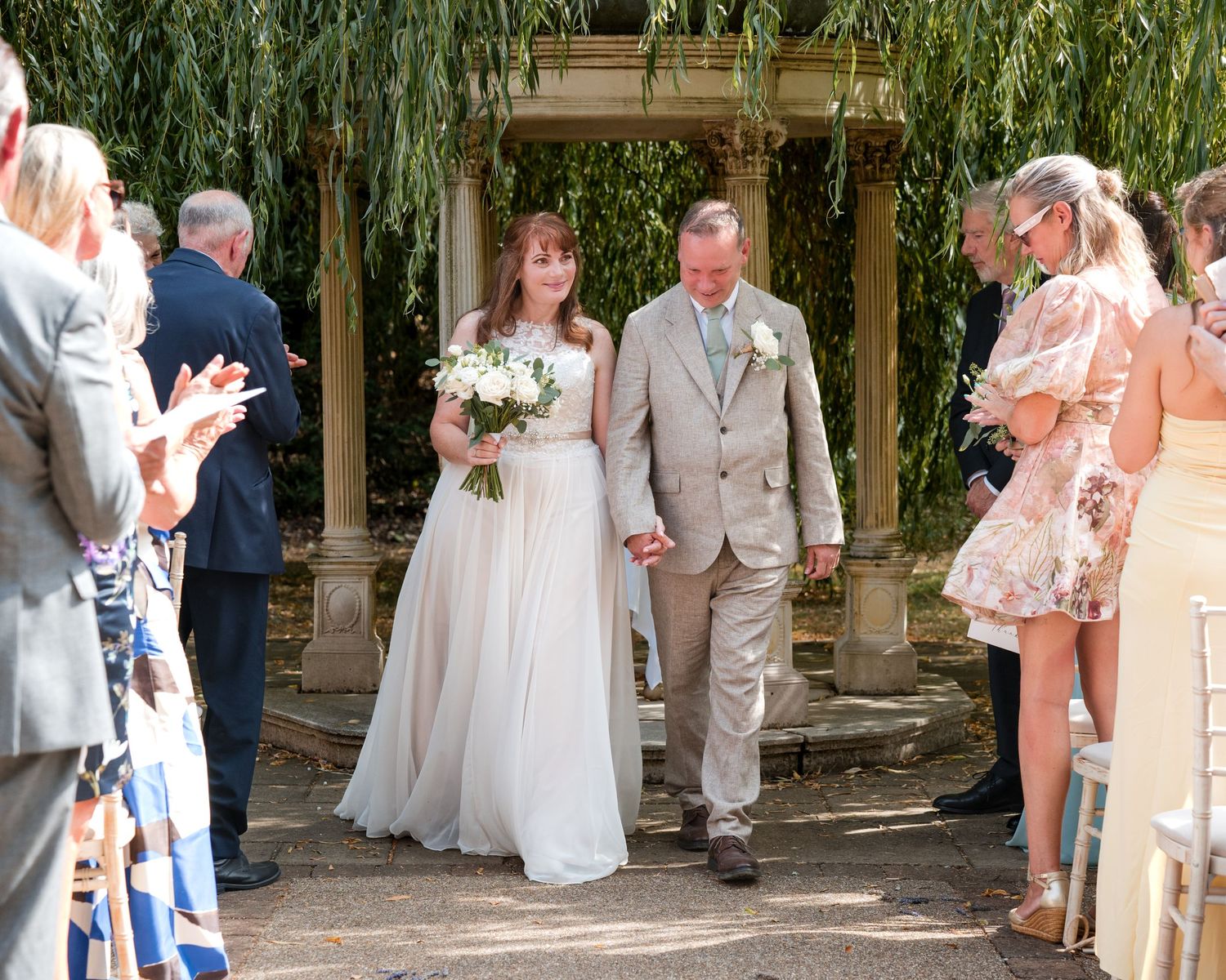 Bride in white gown and groom in tan suit walk down aisle outdoors, guests applauding at elegant garden wedding ceremony.