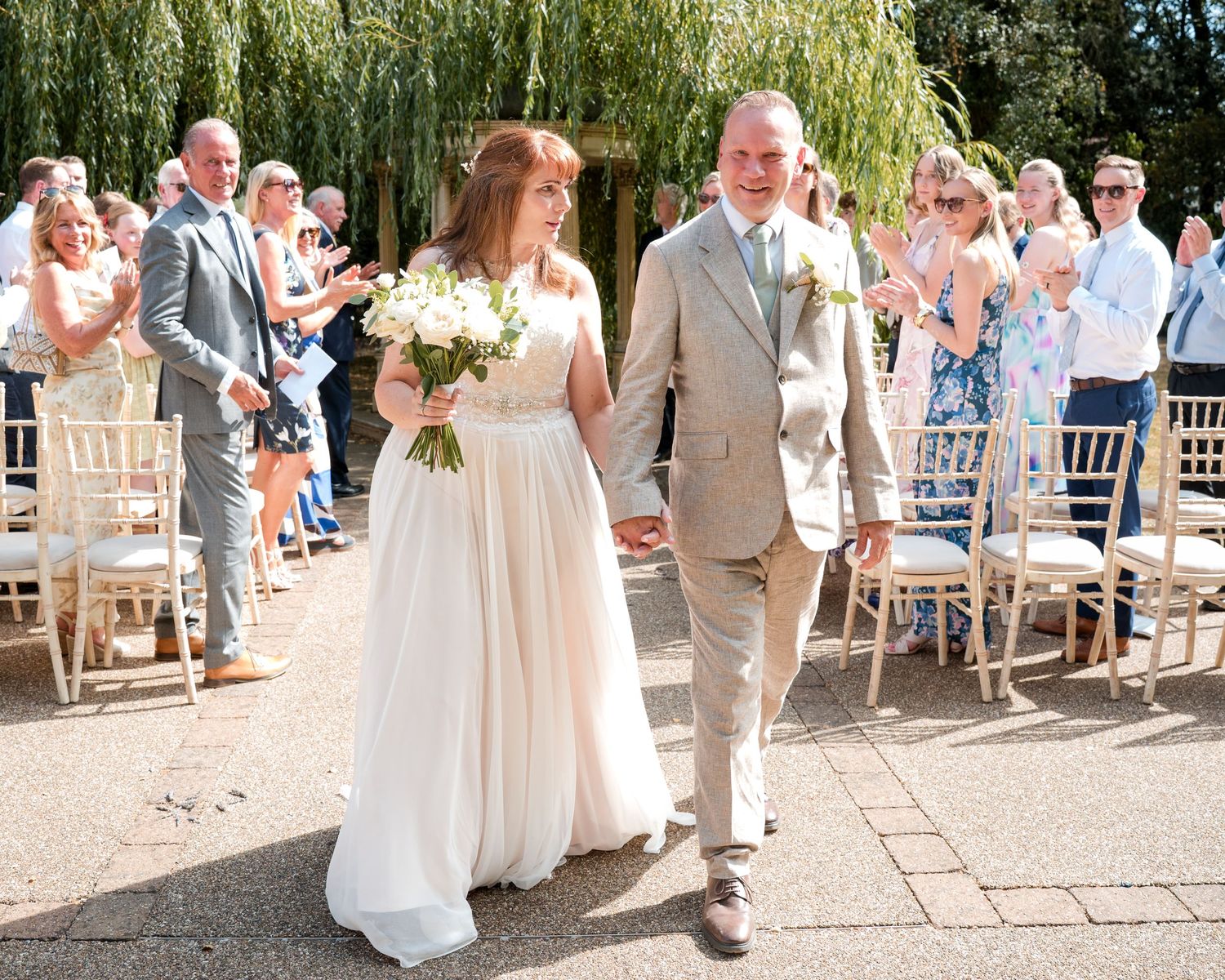 Bride and groom walk down the aisle hand in hand at an outdoor wedding ceremony with guests clapping.