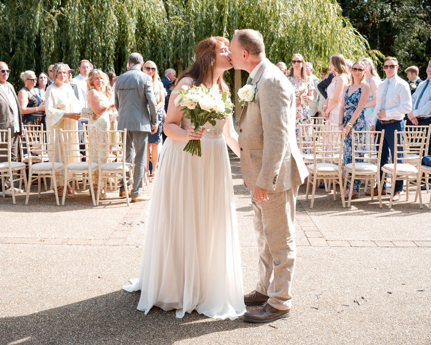 Bride and groom sharing a kiss outdoors at a sunny wedding ceremony with guests and chairs in the background.