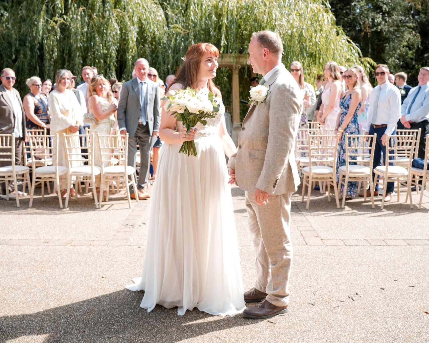 Bride and groom holding hands at outdoor wedding ceremony with guests seated on wooden chairs under willow trees.