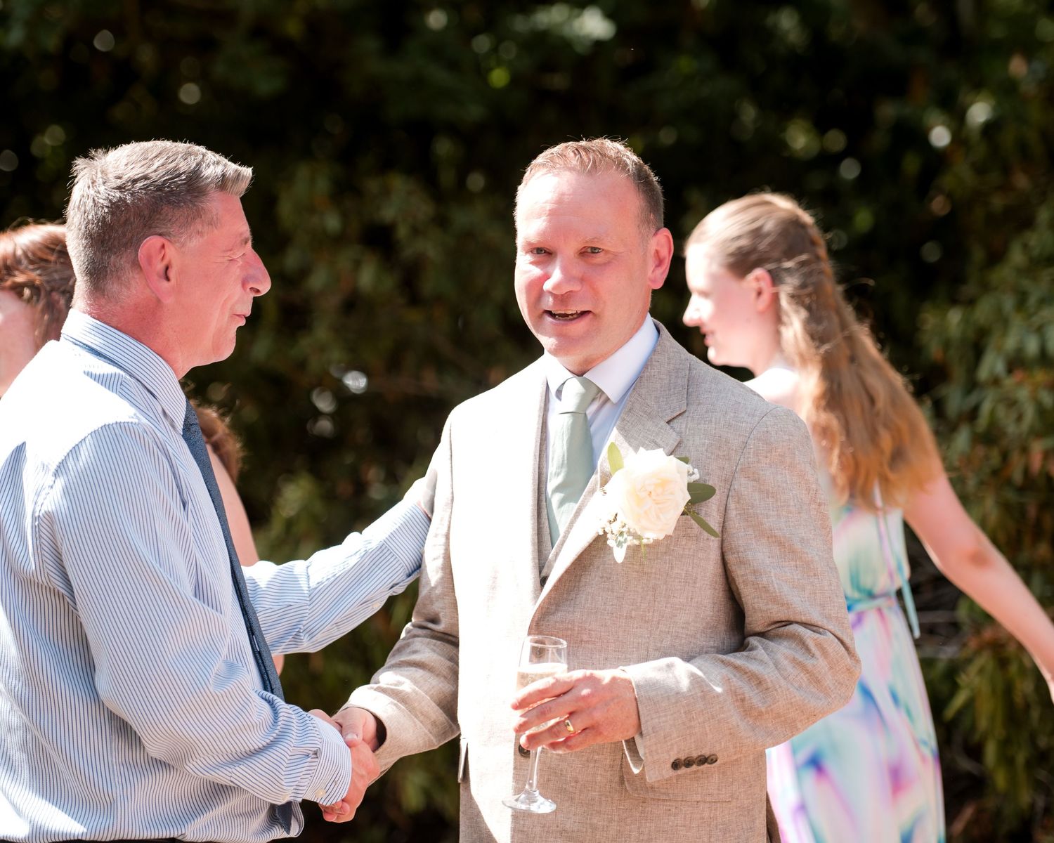 Two men shaking hands at an outdoor wedding reception, one wearing a tan suit with boutonniere holding champagne.