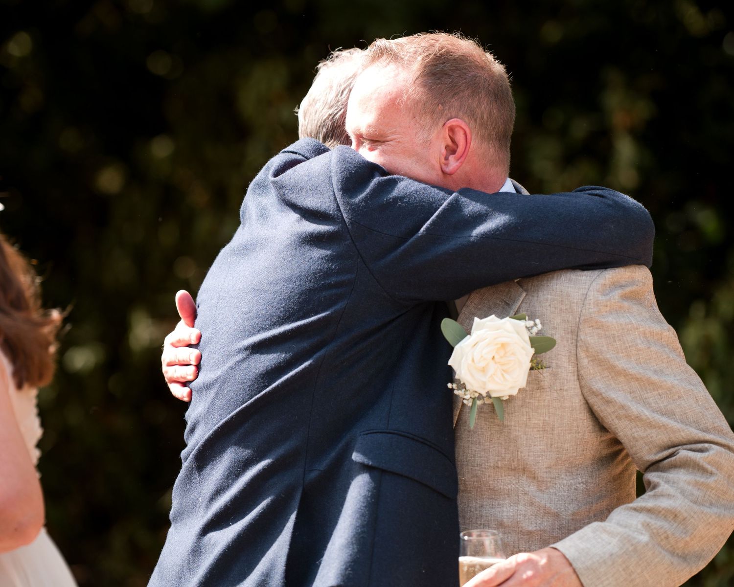 Two men in suits share an emotional hug at an outdoor wedding ceremony, one wearing a white boutonniere.