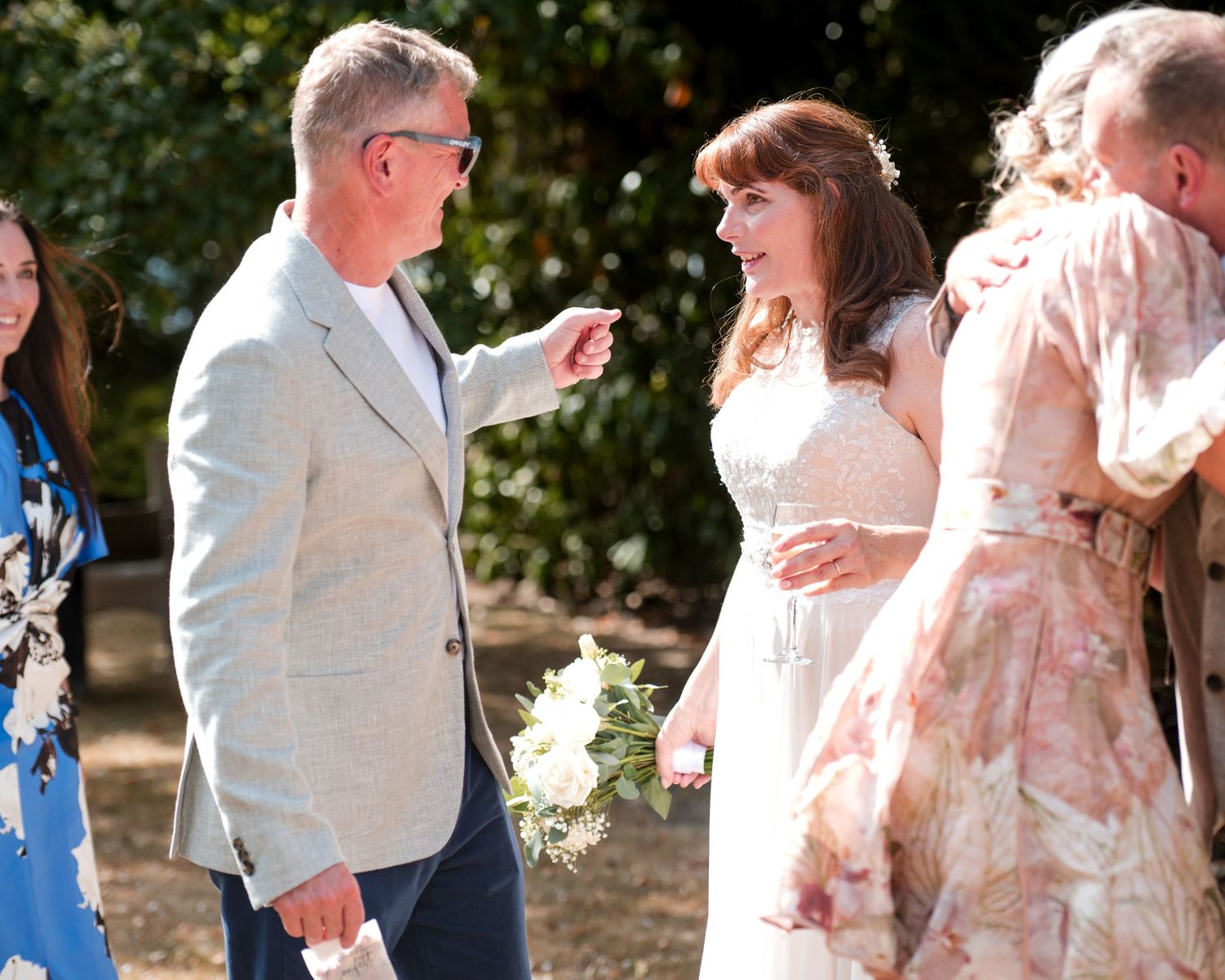 A bride holding flowers converses with a man in a gray suit at an outdoor wedding ceremony.