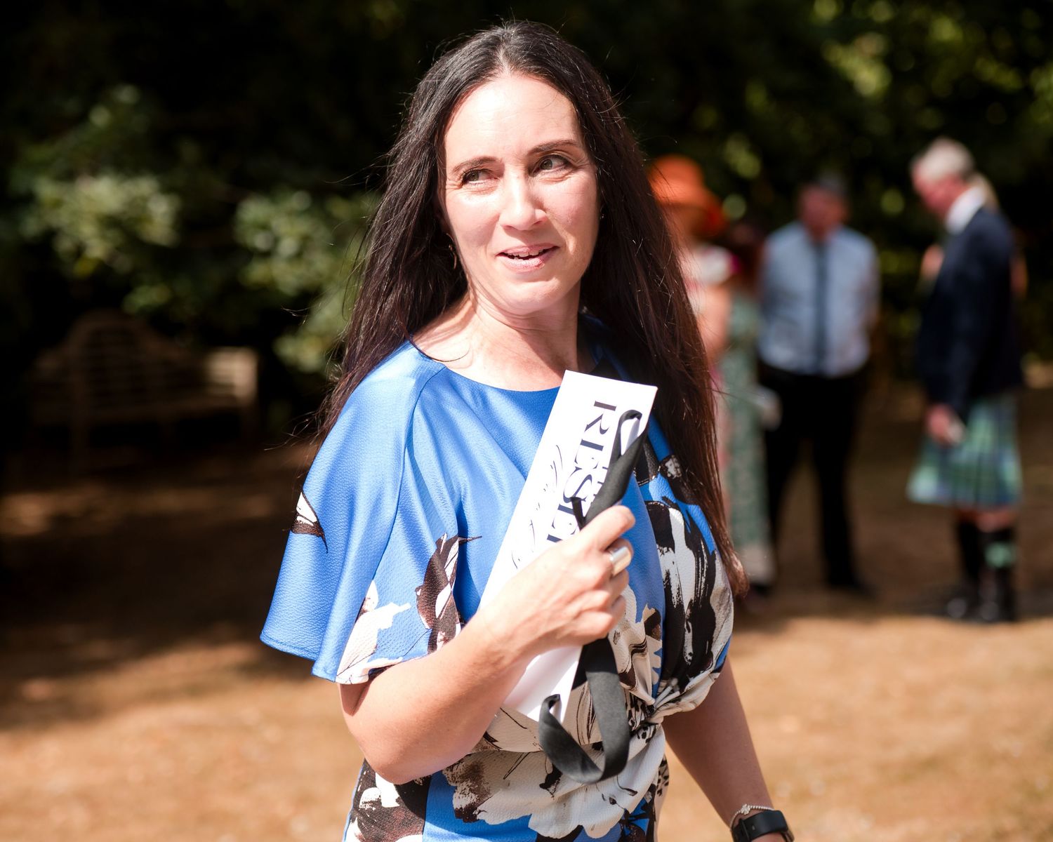Woman with long dark hair in blue floral dress holding a program outdoors at a garden event.