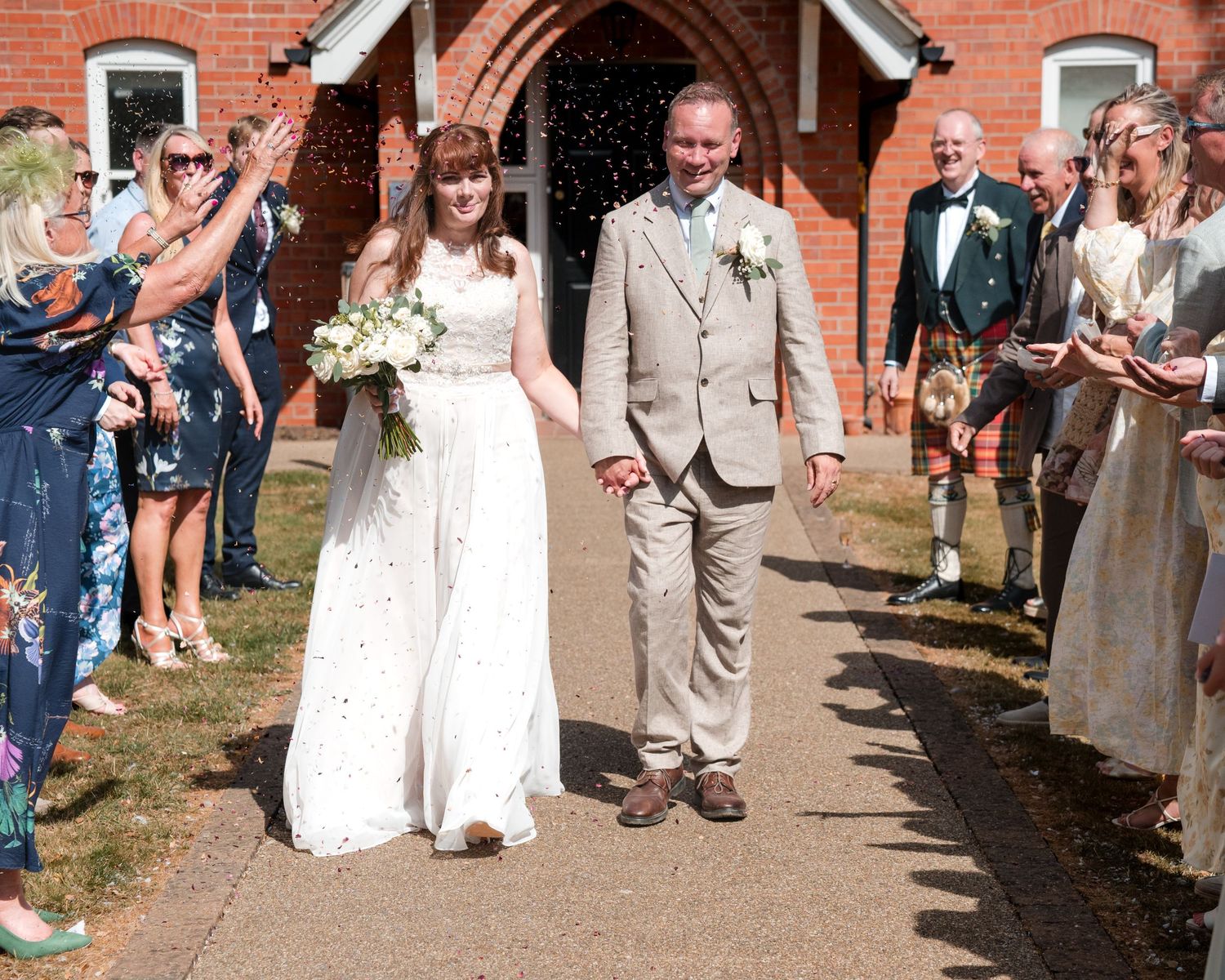 Bride and groom walk hand-in-hand through confetti outside a red brick church on their sunny wedding day.