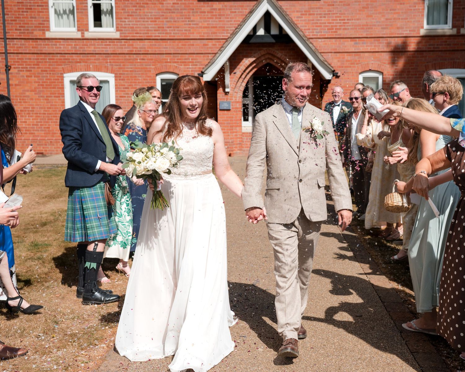 Bride and groom walk through confetti outside red brick church surrounded by smiling wedding guests on sunny day.