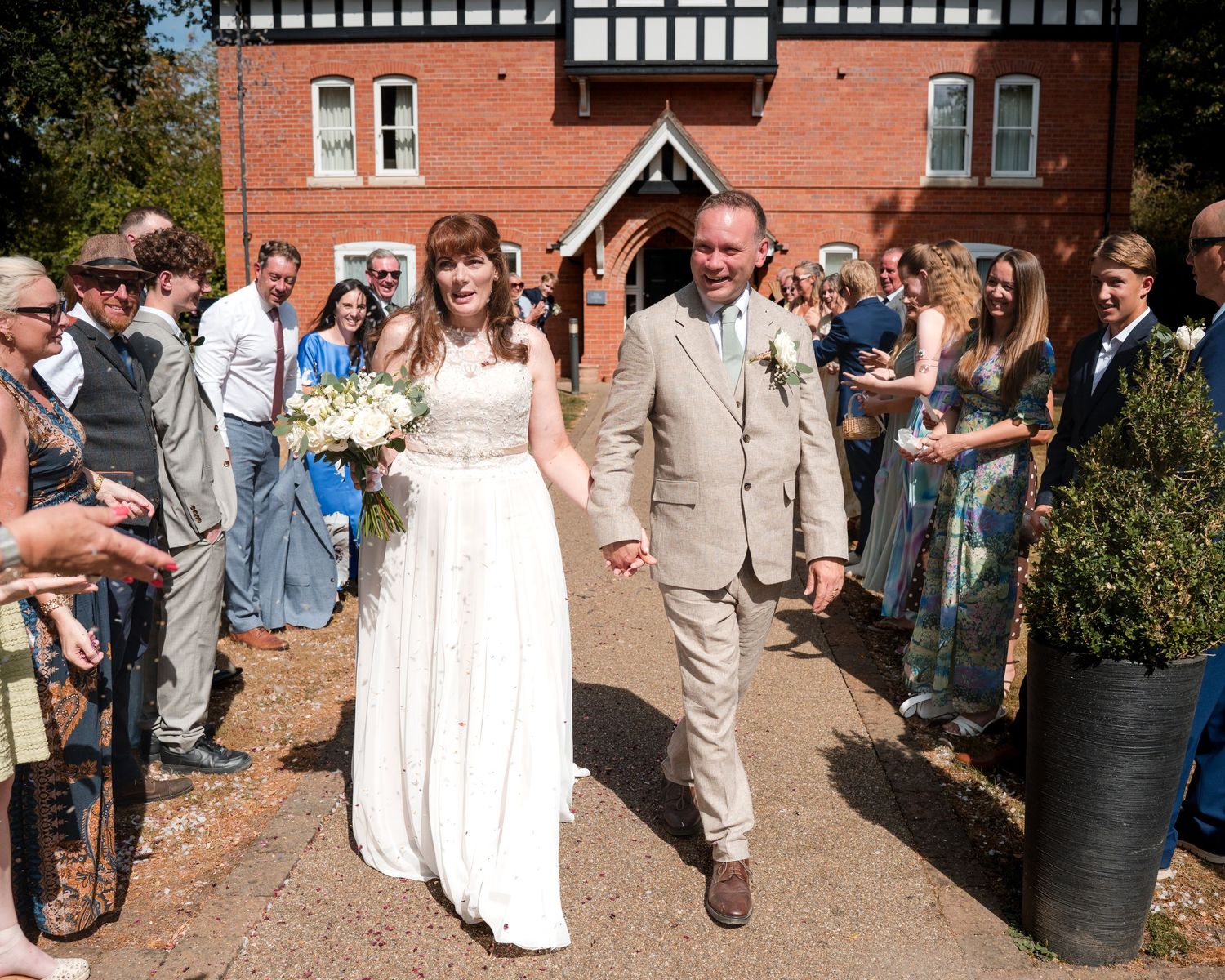 Bride and groom walk hand-in-hand past cheering guests outside a red brick building on their wedding day.