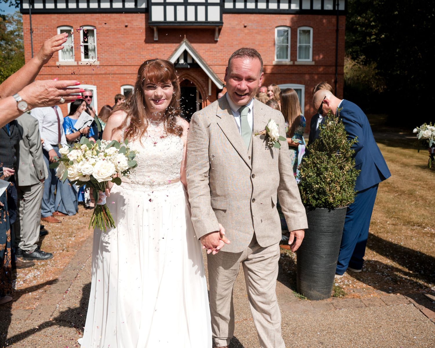 Bride and groom walk hand in hand outside a red brick venue, surrounded by wedding guests on a sunny day.