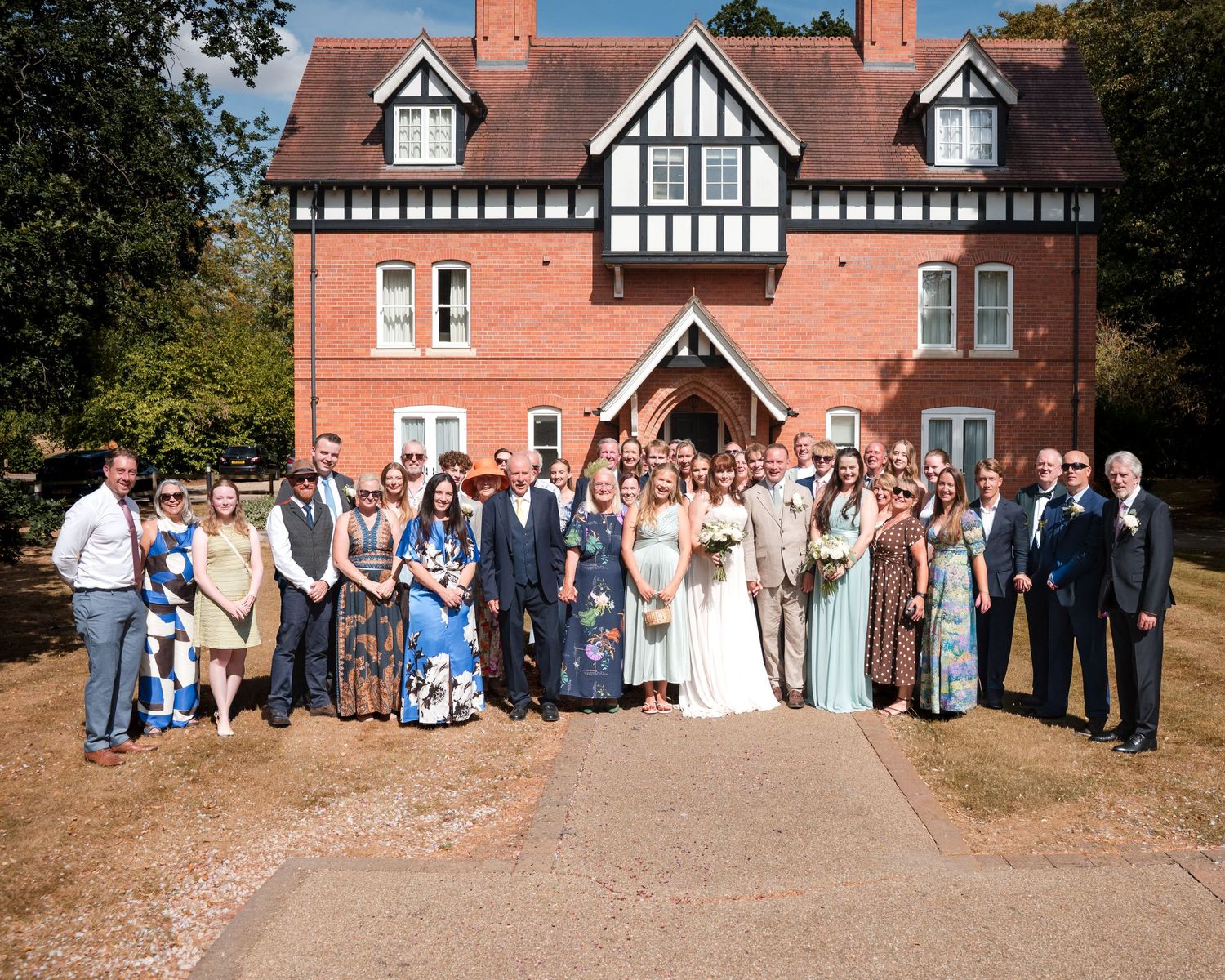 Wedding party group photo outside a red brick Tudor-style manor house on a sunny day.