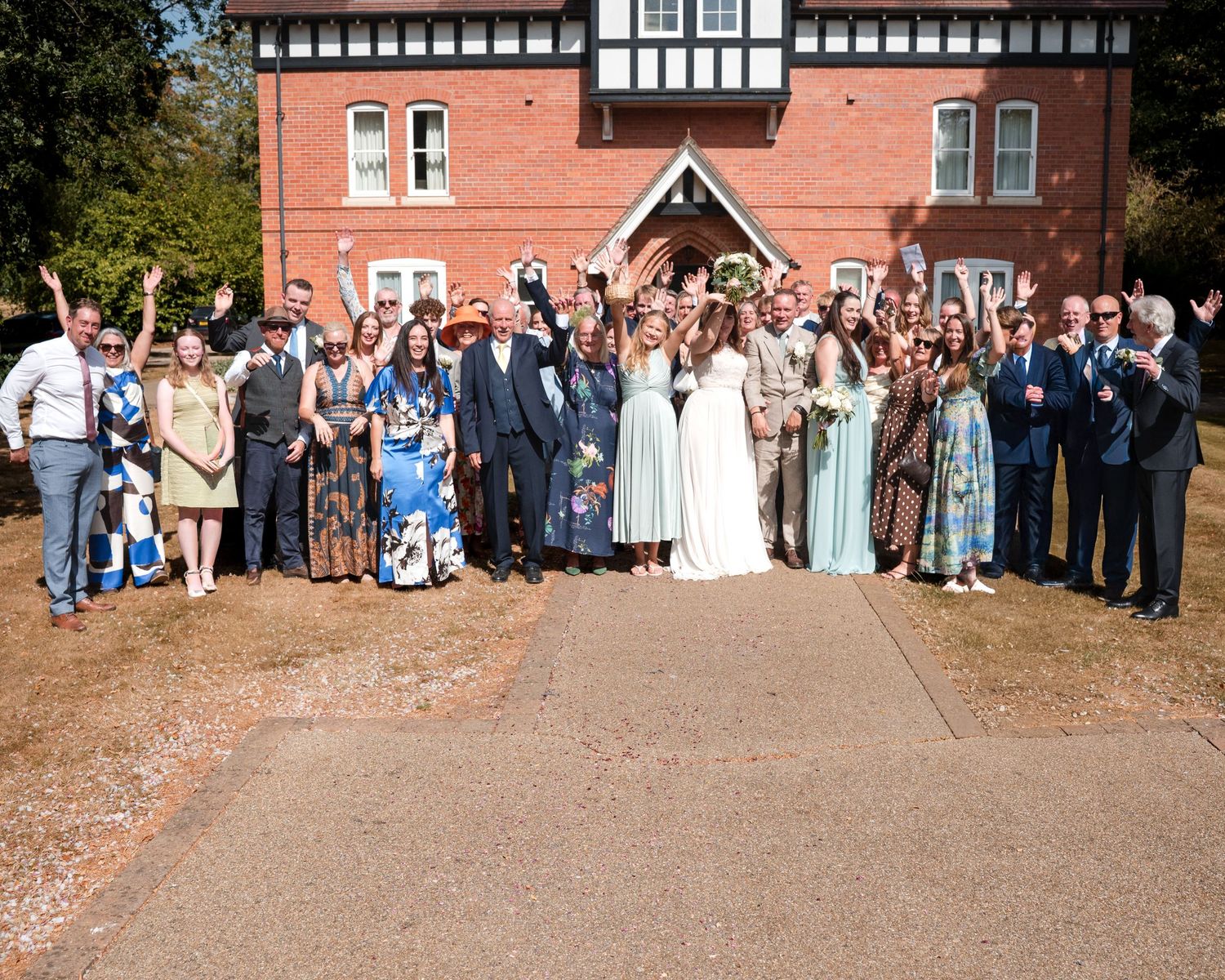 Large wedding party posing joyfully outside a red brick Tudor-style building on a sunny day.
