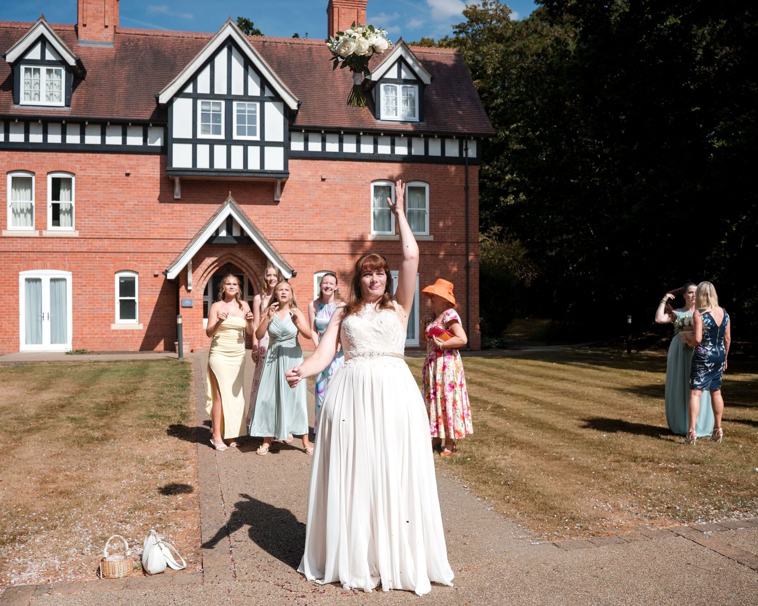 Bride tossing bouquet outside a red brick Tudor manor house with bridesmaids watching in colorful dresses.