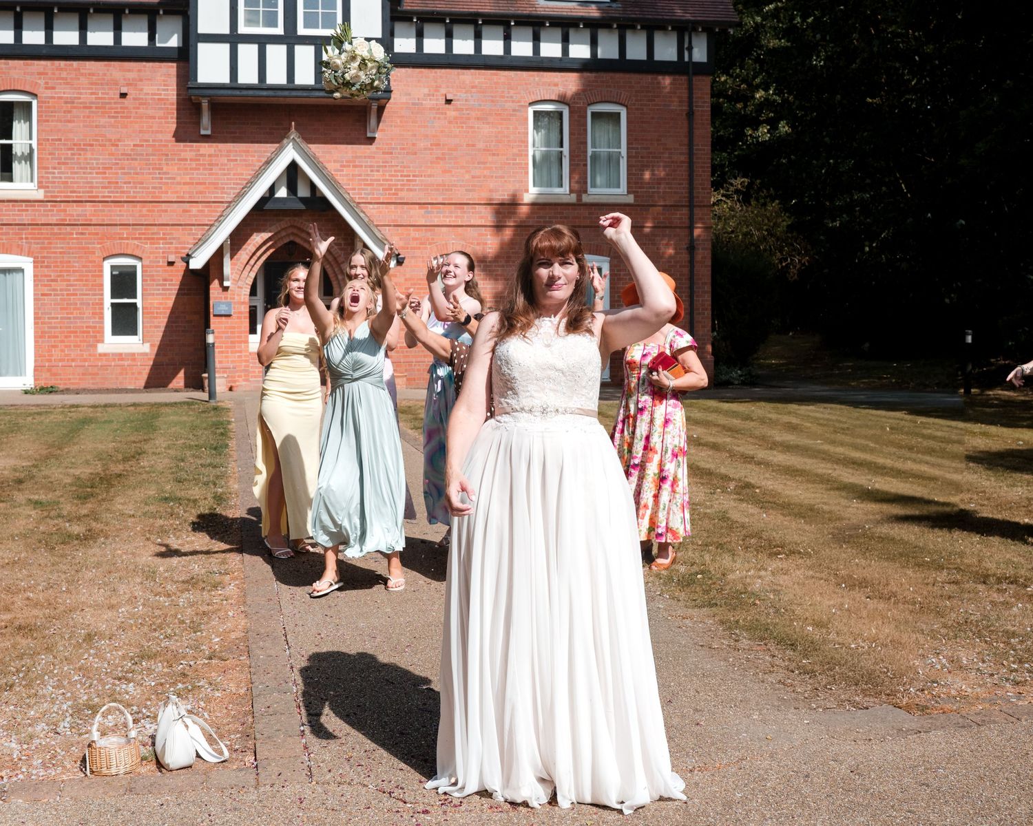 Bride in white gown tosses bouquet to bridesmaids in colorful dresses outside a red brick building.