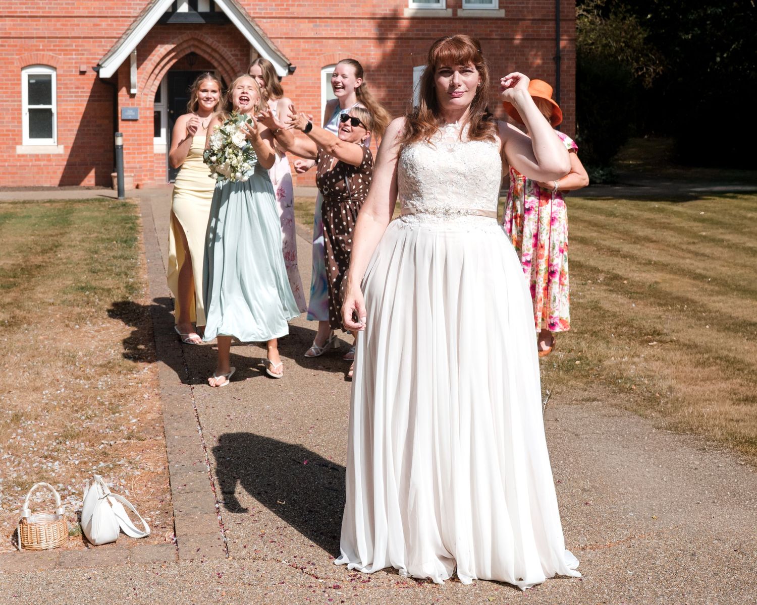 Bride in white gown tosses bouquet to excited bridesmaids outside red brick church.