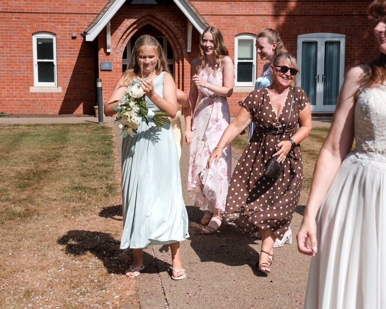 Bridesmaids in pastel dresses laughing outside a red brick church on a sunny wedding day.