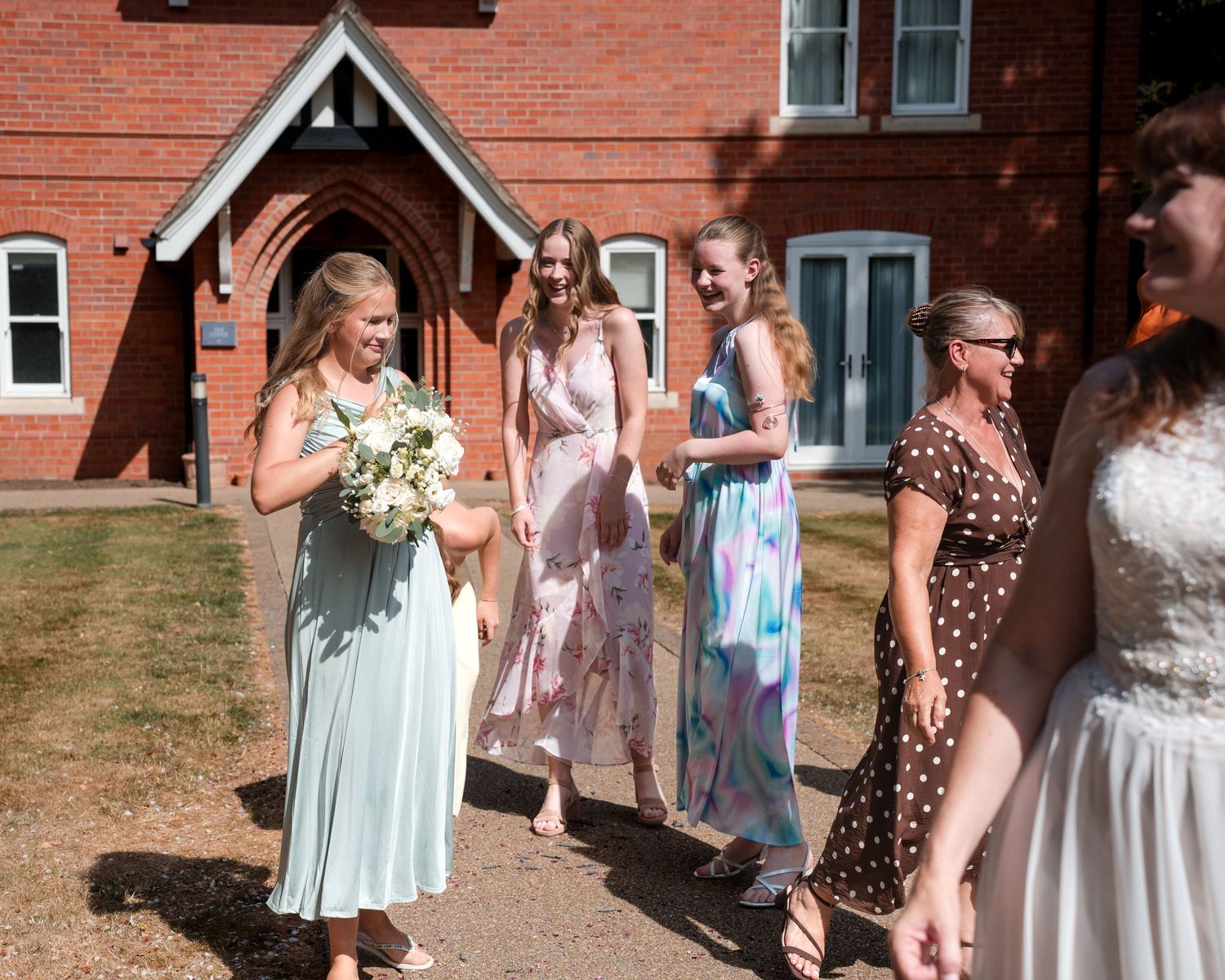 Bridesmaids in pastel dresses walk outside a red brick church on a sunny wedding day.