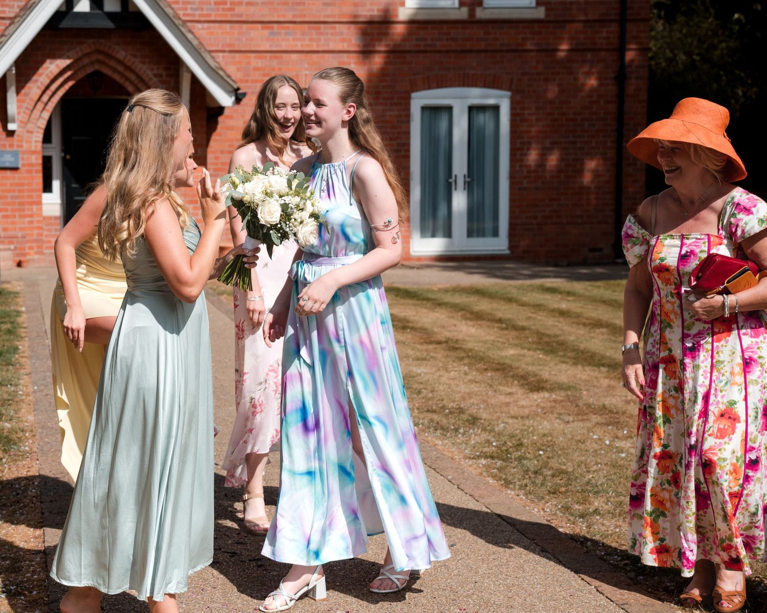 Young women in elegant dresses gather outdoors near a brick church, one holding a bouquet, as an older woman in orange hat looks on.