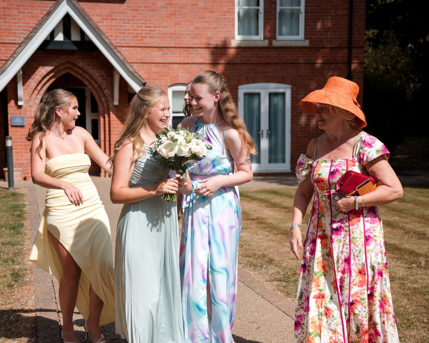 Three bridesmaids in pastel gowns hold white flowers outside a red brick church, greeted by a woman in floral dress.