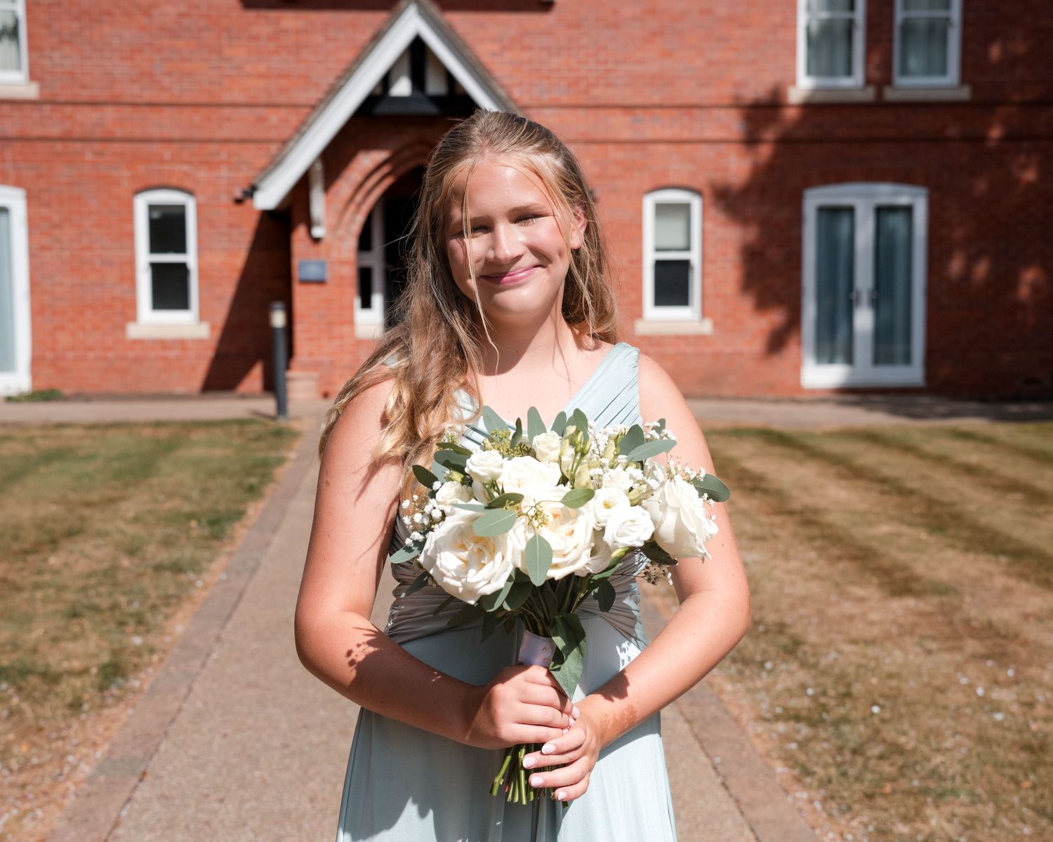 Young girl in light blue dress holding white flower bouquet stands smiling outside a red brick building.
