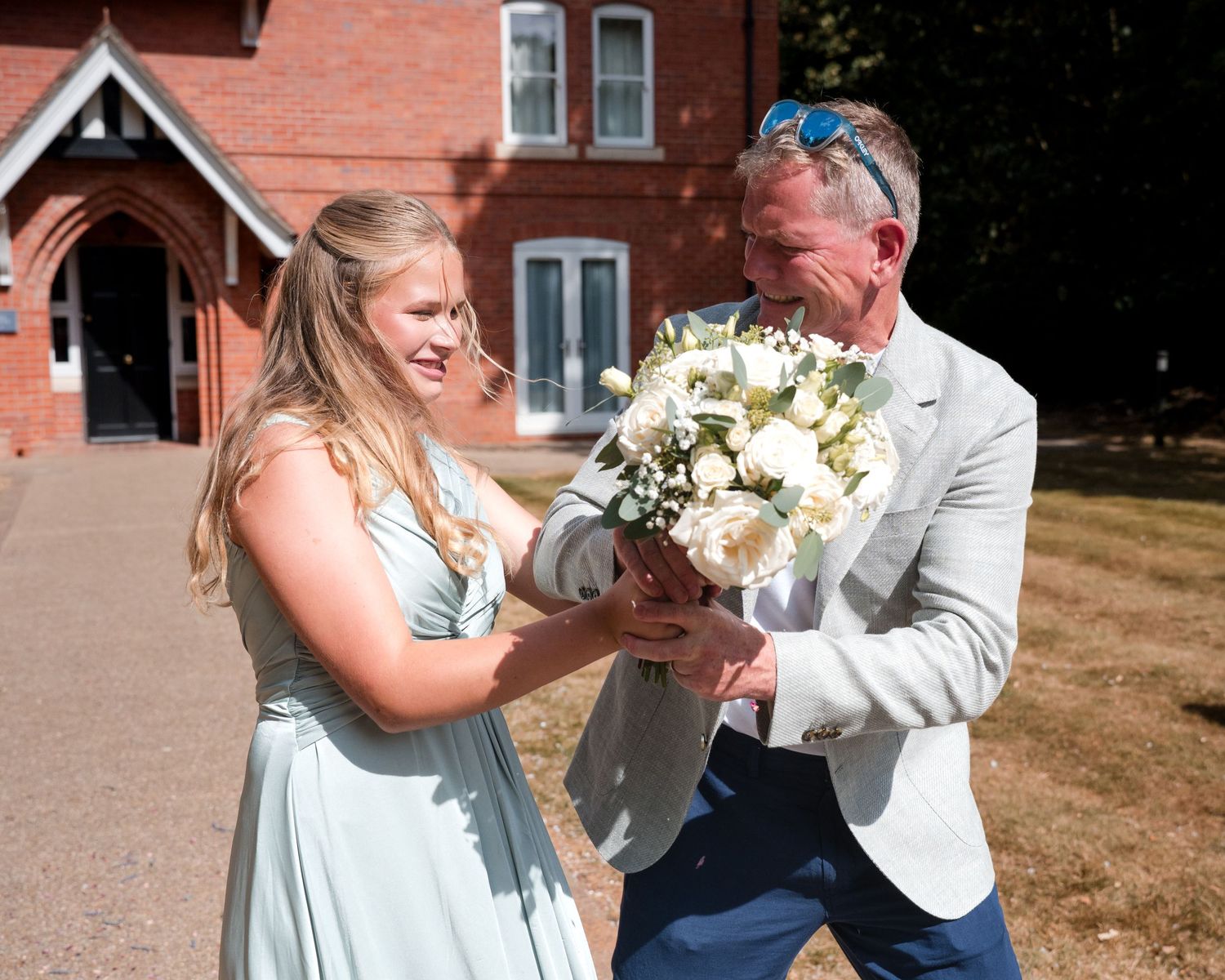 Bride in mint dress and groom in grey suit smiling while holding white floral bouquet outside red brick building.