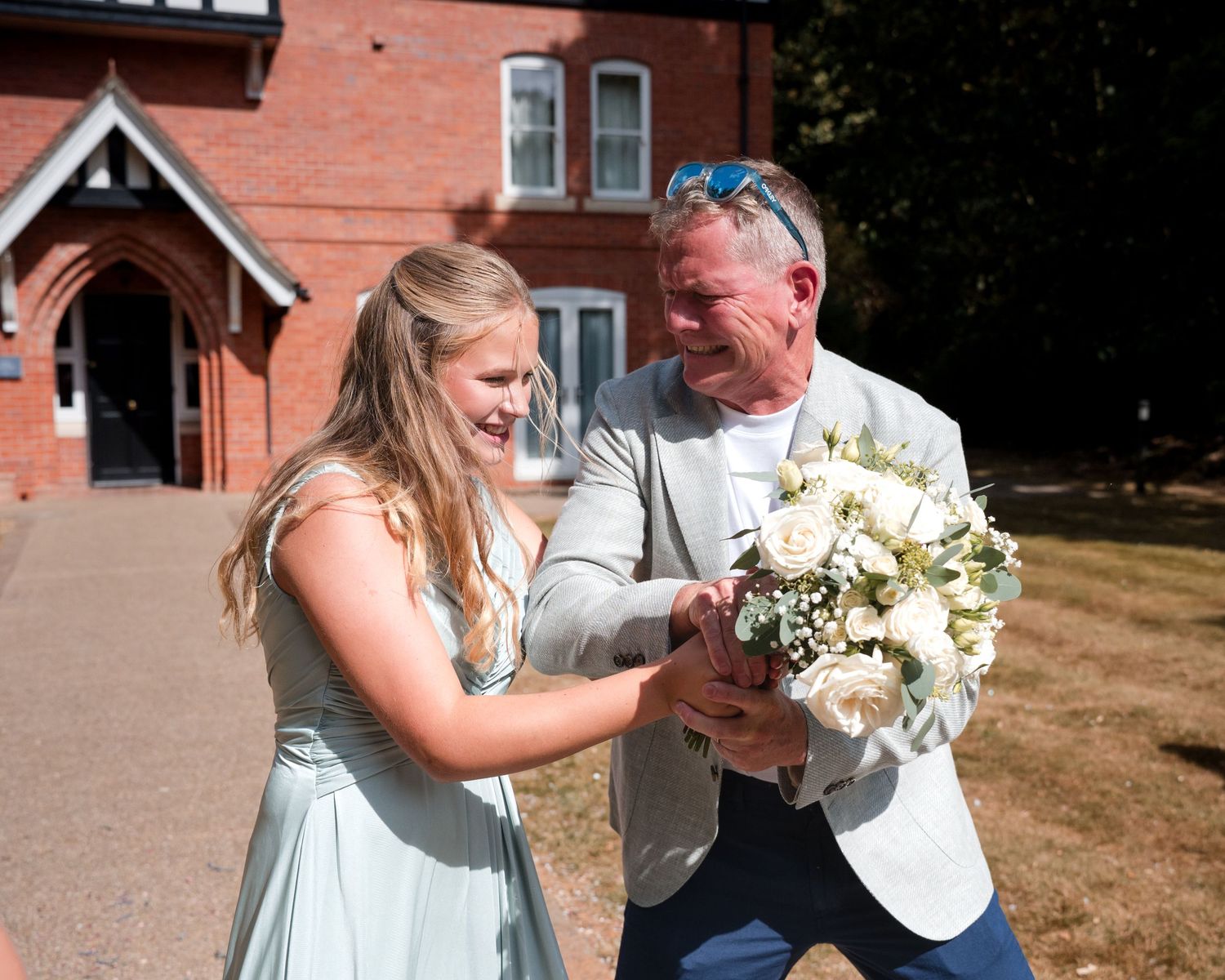 Bride in mint dress laughing with older man holding white rose bouquet outside red brick wedding venue.