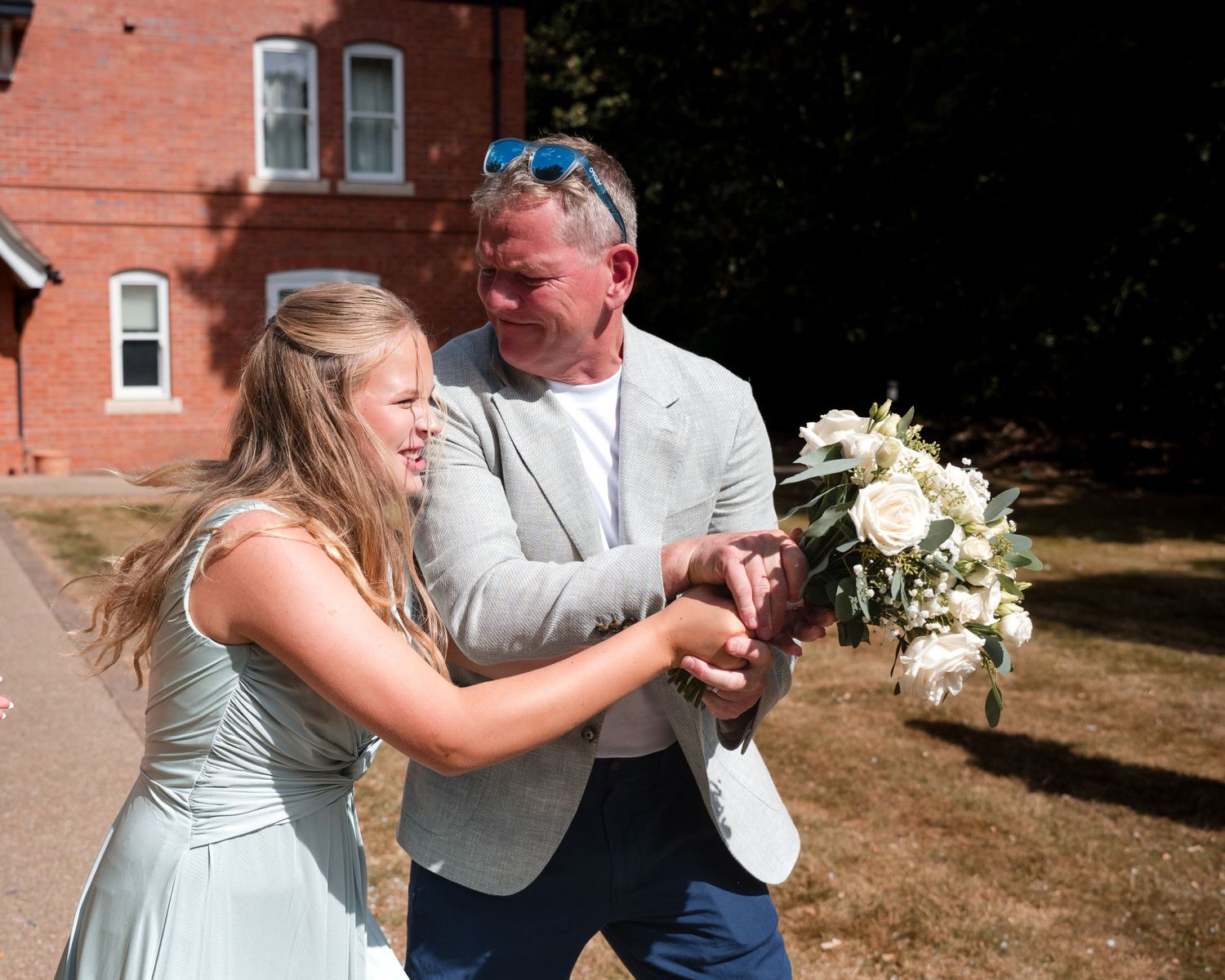 Bride in mint dress and groom in grey suit share a joyful moment with white bouquet outside red brick building.