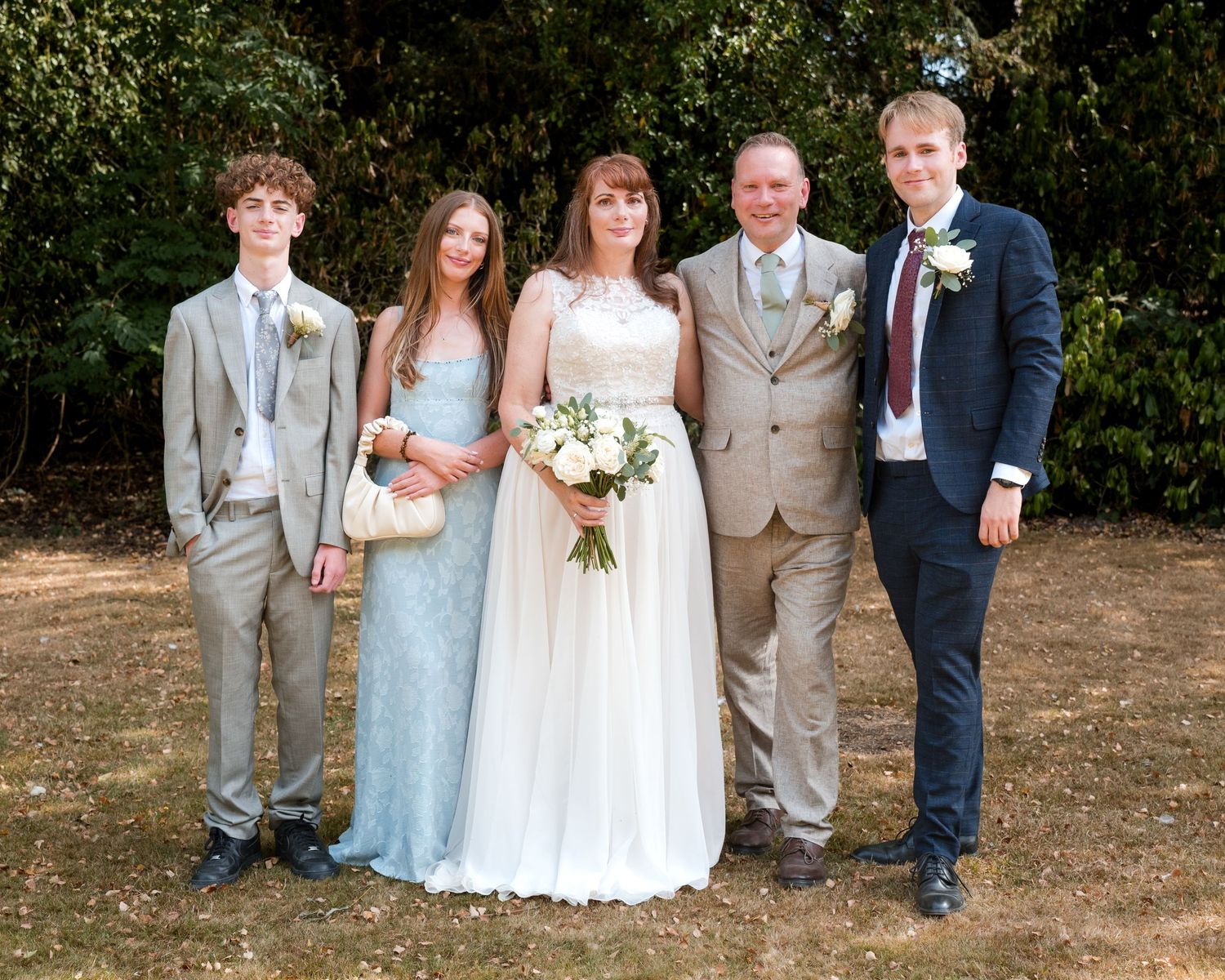 Wedding party of five posing outdoors, bride in white gown holding bouquet, flanked by guests in formal attire.