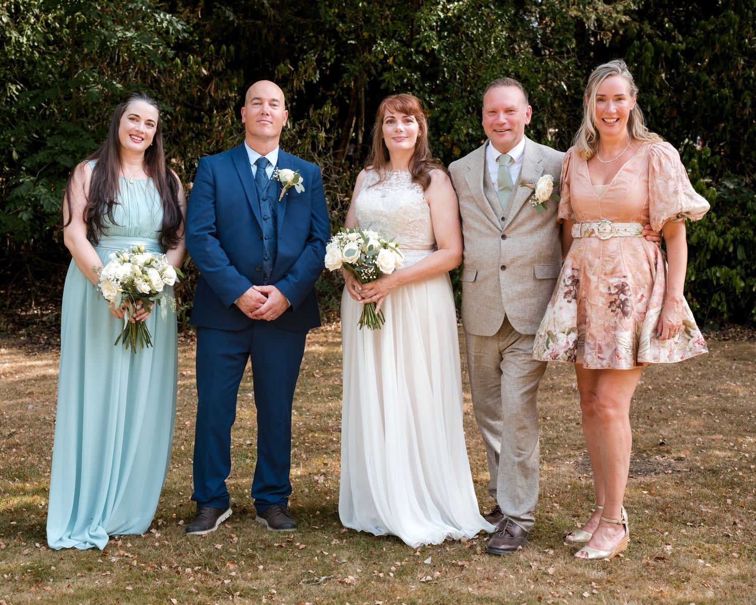 Wedding party of five posing outdoors, bride in white gown, groom in tan suit, bridesmaids in blue and pink dresses.