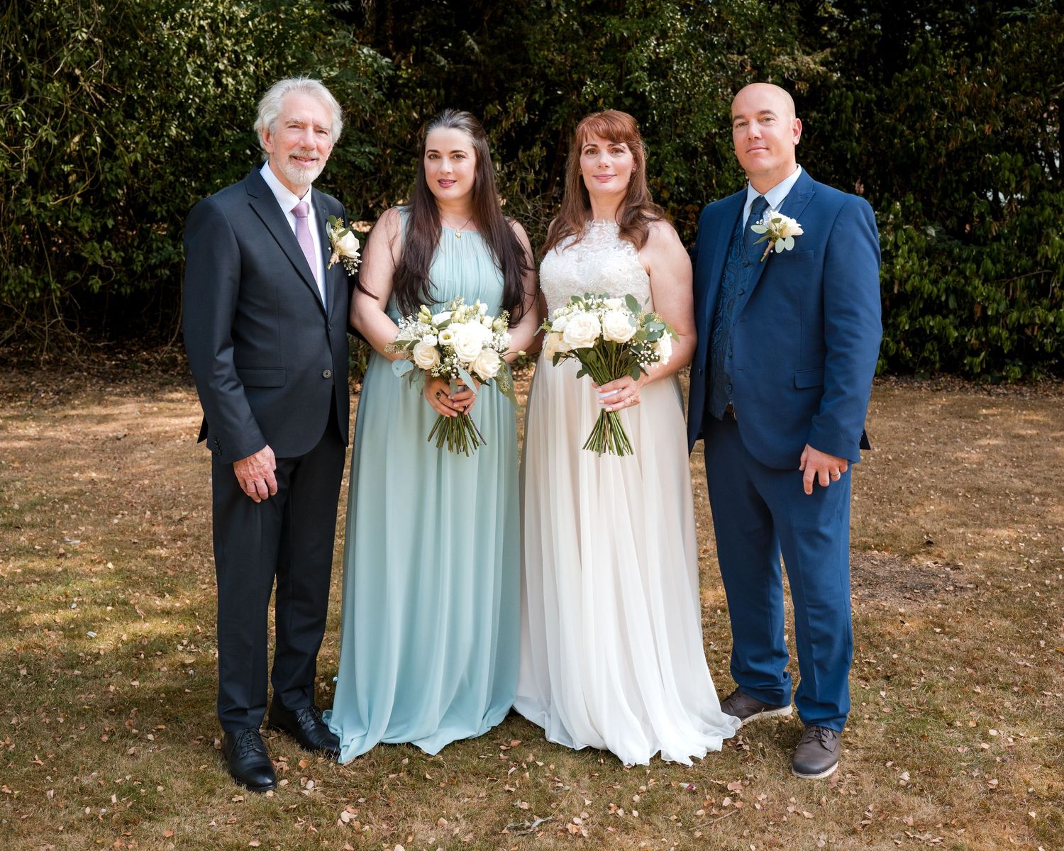 Wedding party posing outdoors, bride in white gown with bouquet, bridesmaid in blue dress, two men in suits.
