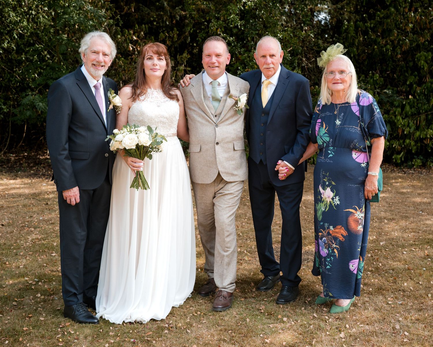 Wedding group photo with bride in white gown holding bouquet, groom, and three guests posing outdoors.