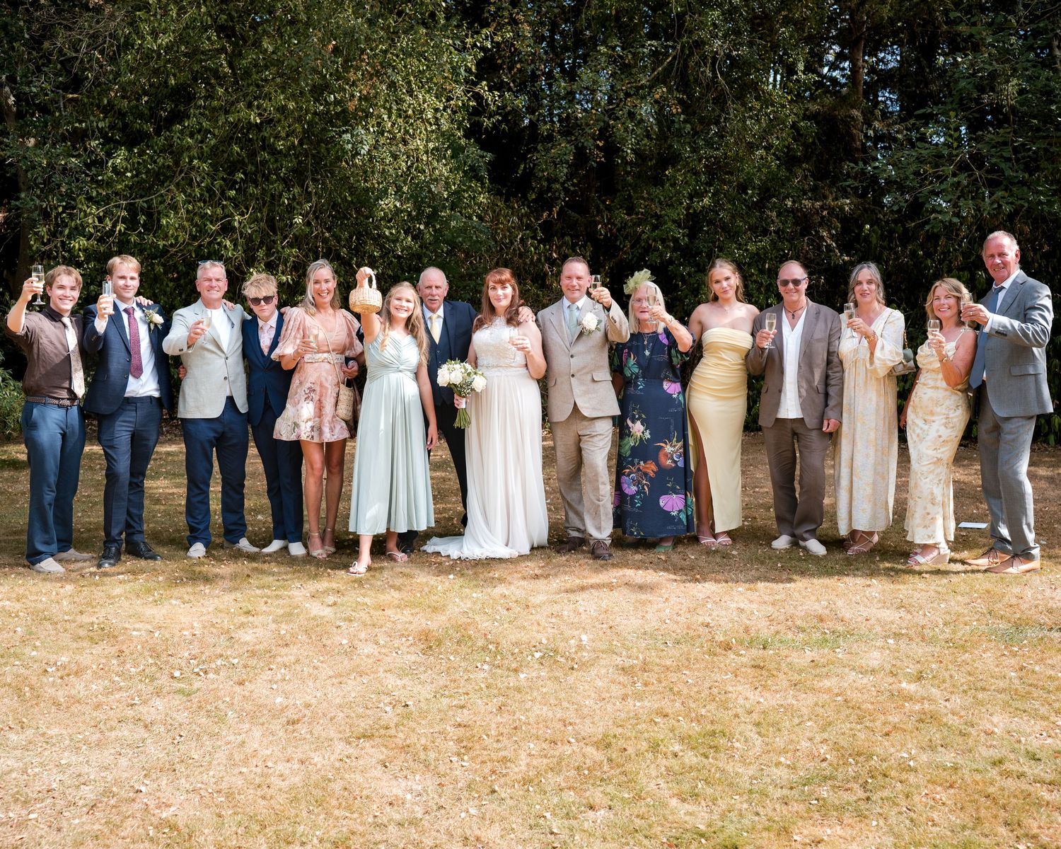 Wedding party celebrating outdoors on sunny day, bride and groom surrounded by guests raising glasses in a toast.