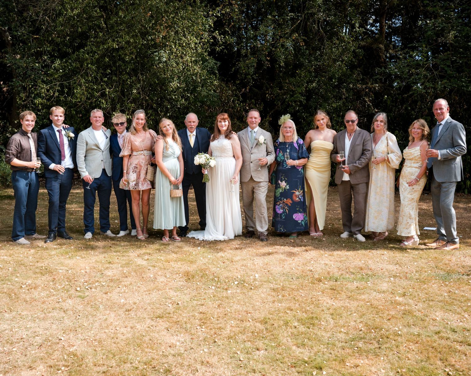 Large wedding party group photo outdoors on sunny day, bride and groom surrounded by family and friends on green lawn.